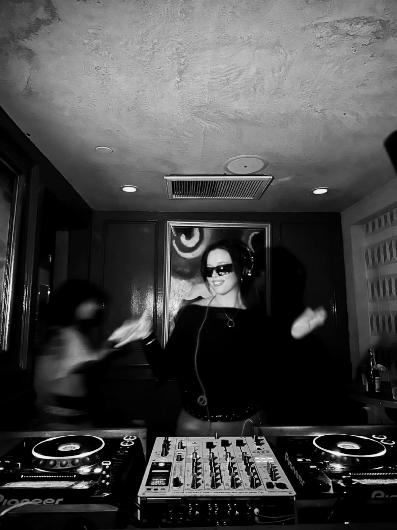 A woman DJ wearing sunglasses and headphones, standing behind DJ equipment, smiling and dancing in a dimly lit room with a portrait of a woman in the background.