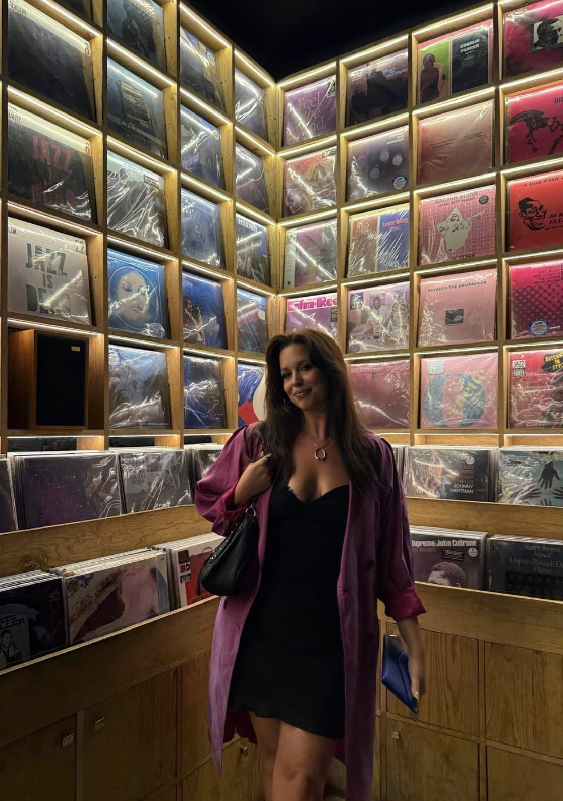 A woman standing inside a record store surrounded by shelves of vinyl records, with a mix of pink and purple album covers visible behind her.