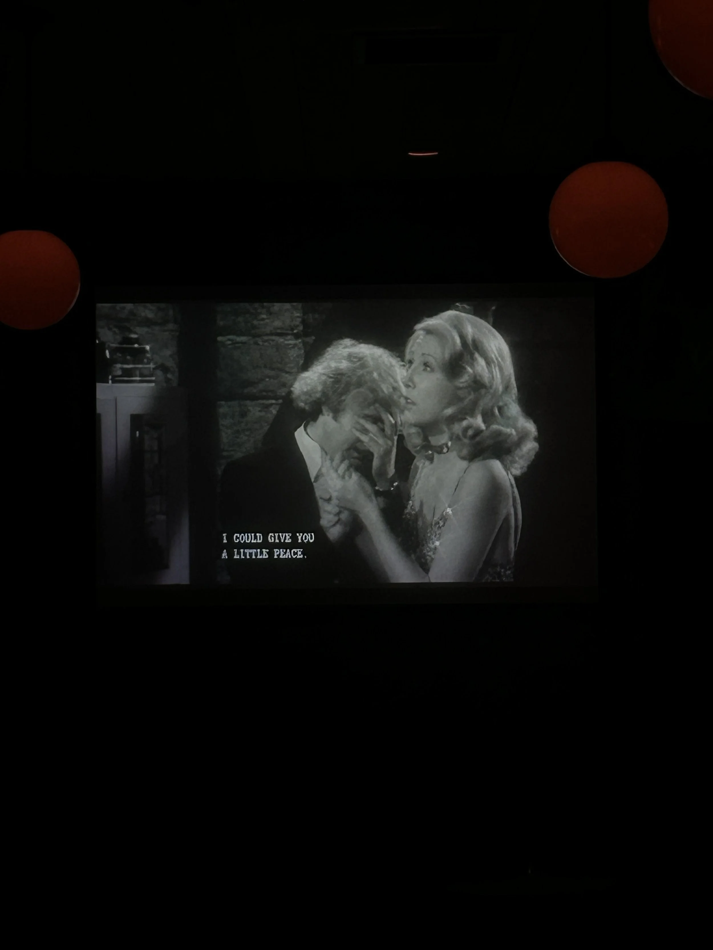 A scene from a black-and-white movie featuring a man and woman sharing a kiss. The woman has wavy blonde hair and is wearing a dress, while the man has short light-colored hair and is in a suit. Text at the bottom reads, "I could give you a little peace."