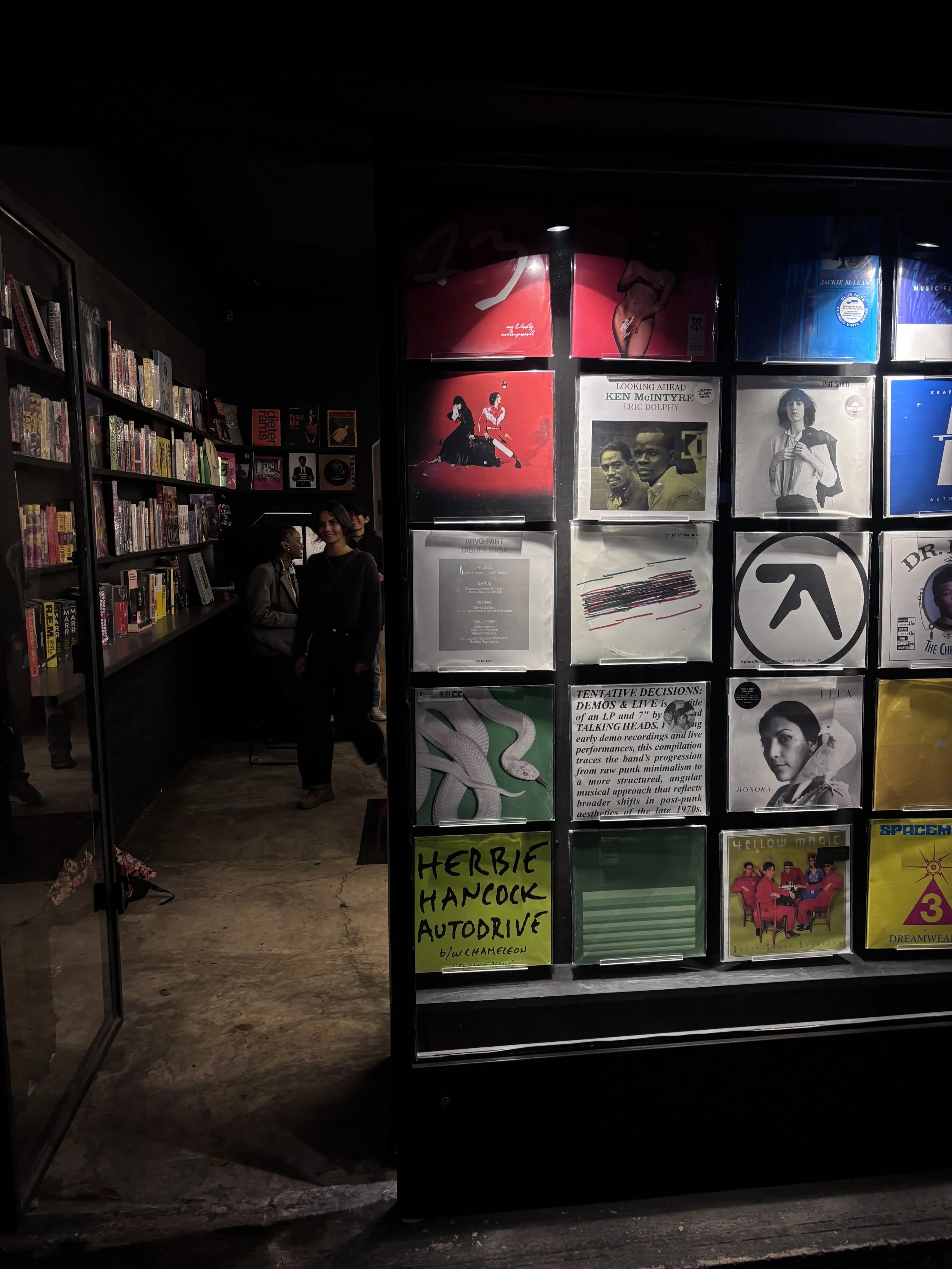 A dimly lit record store with shelves of vinyl records on the left, and a display of album covers on the right, including various album artwork and some posters. There are several people, including two women standing near the entrance, visible in the background.