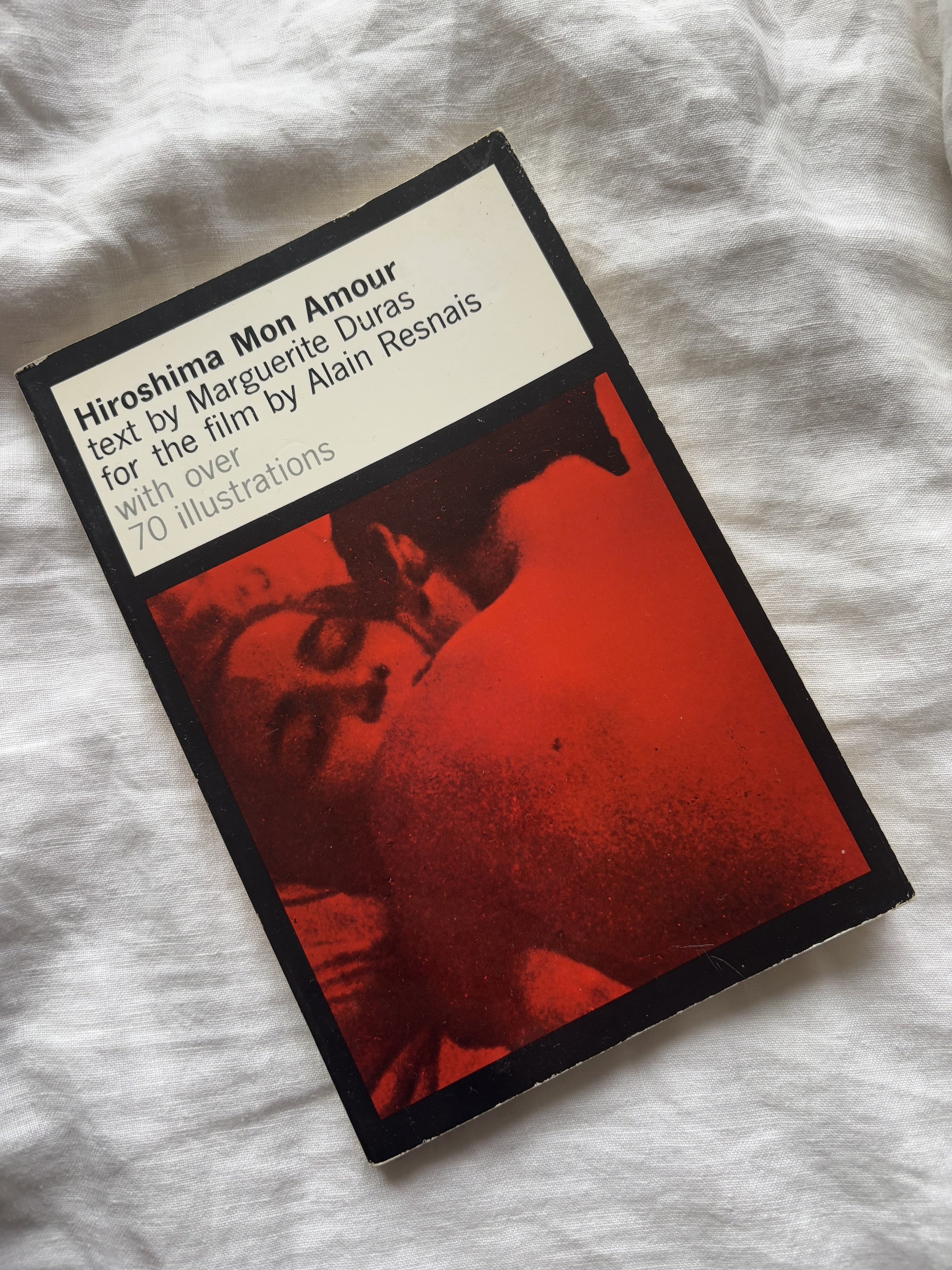Close-up of a book cover titled 'Hiroshima Mon Amour' with text about a film by Alain Resnais, featuring a red and black graphic image, placed on a crumpled white fabric surface.