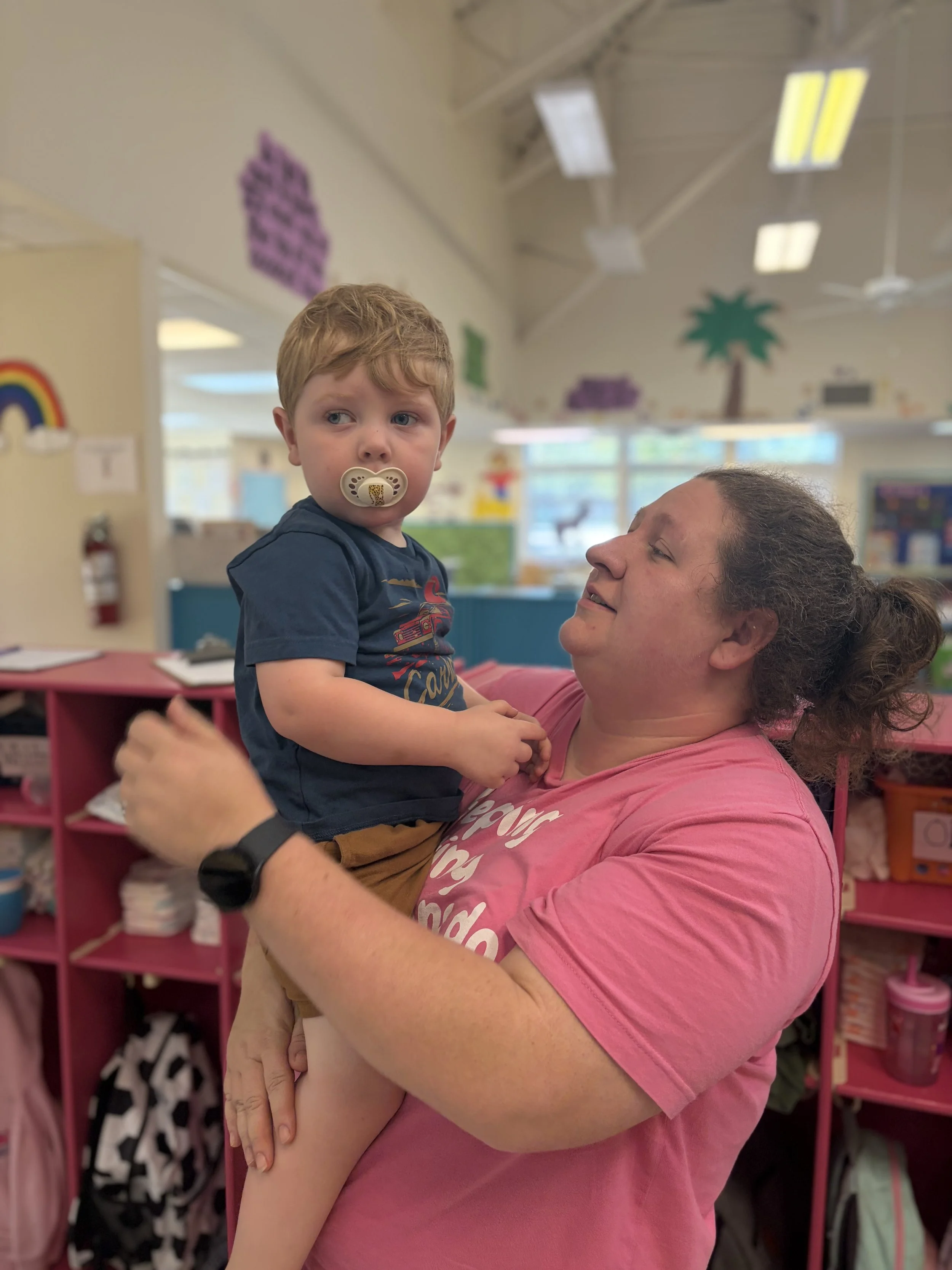 A woman with curly hair smiling while holding a young boy with a pacifier in his mouth, in a colorful indoor setting, possibly a classroom or daycare.