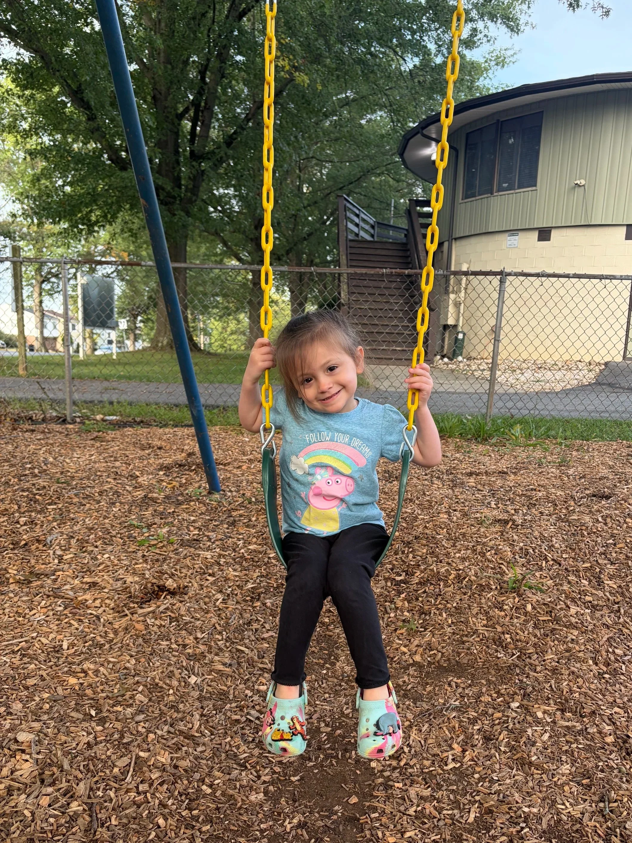 A smiling young girl sitting on a swing at a playground, wearing a Peppa Pig T-shirt, black pants, and colorful shoes with animal designs, surrounded by wood chips and trees.