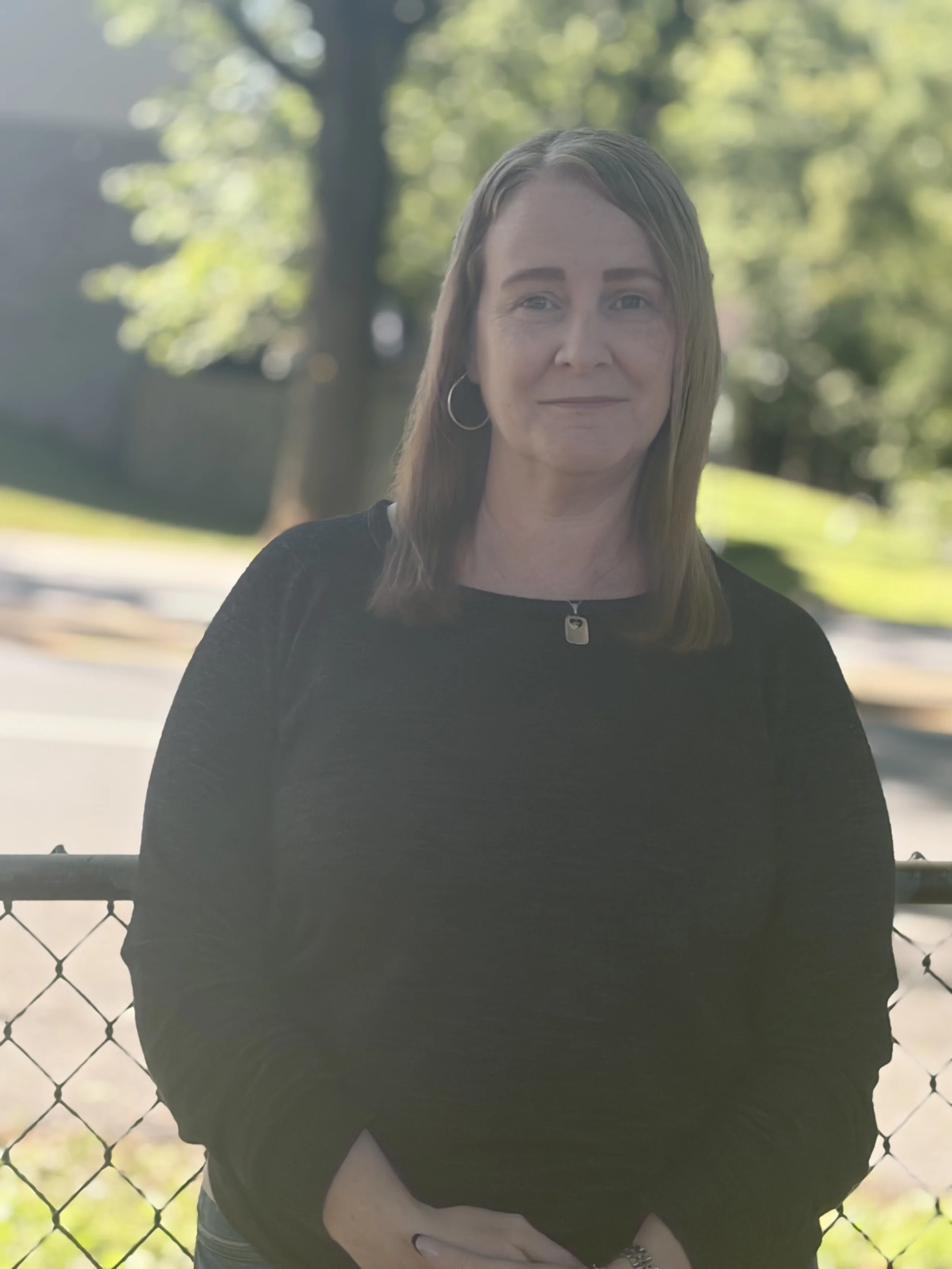 A woman with light skin, shoulder-length brown hair, wearing hoop earrings and a black top, standing outside in front of a chain-link fence with trees and sunlight in the background.