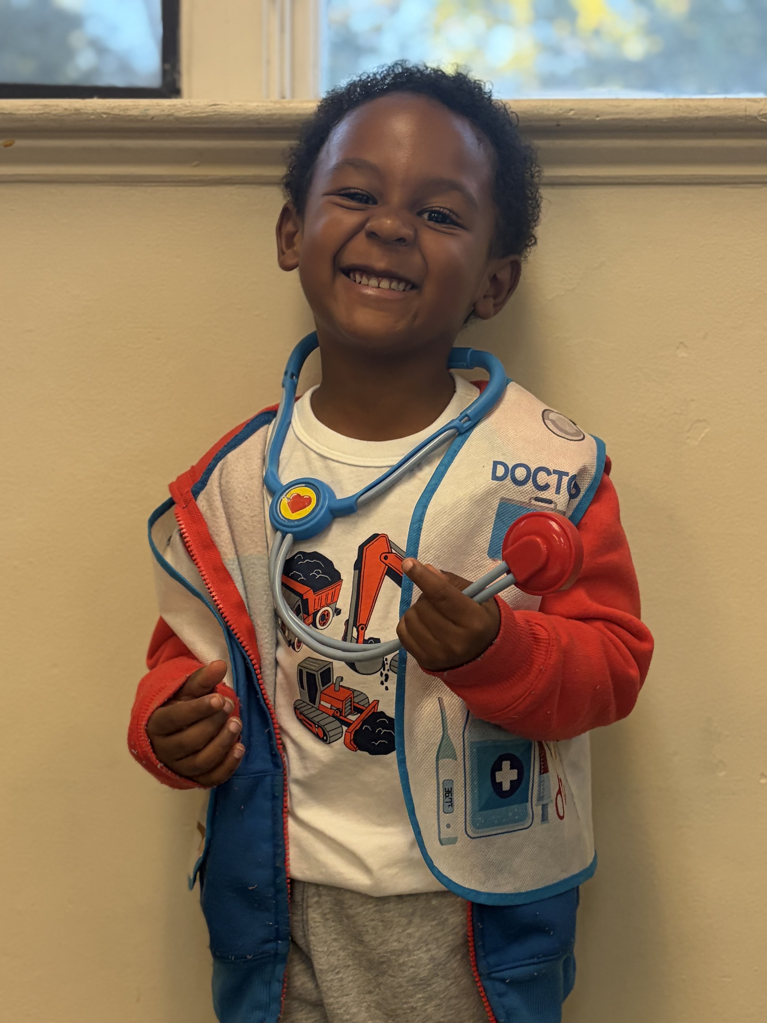 A young boy with dark curly hair smiling and pointing at himself, wearing a toy stethoscope around his neck, a white graphic t-shirt, and a colorful vest with a nurse or doctor theme, standing against a beige wall near a window with a blurred outdoor view.