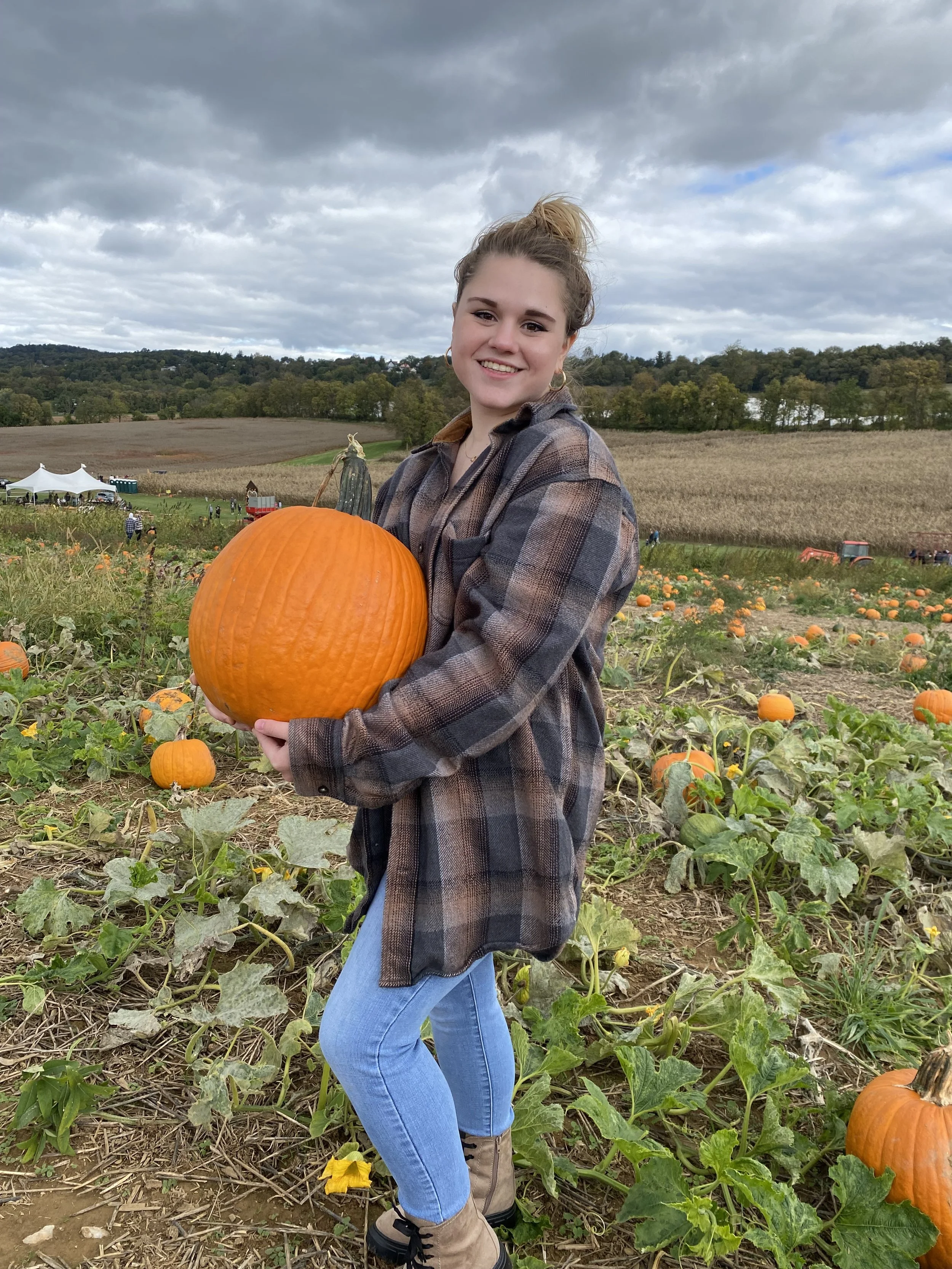 A young woman holding a large orange pumpkin in a pumpkin patch on a cloudy day, with pumpkins and farm structures in the background.