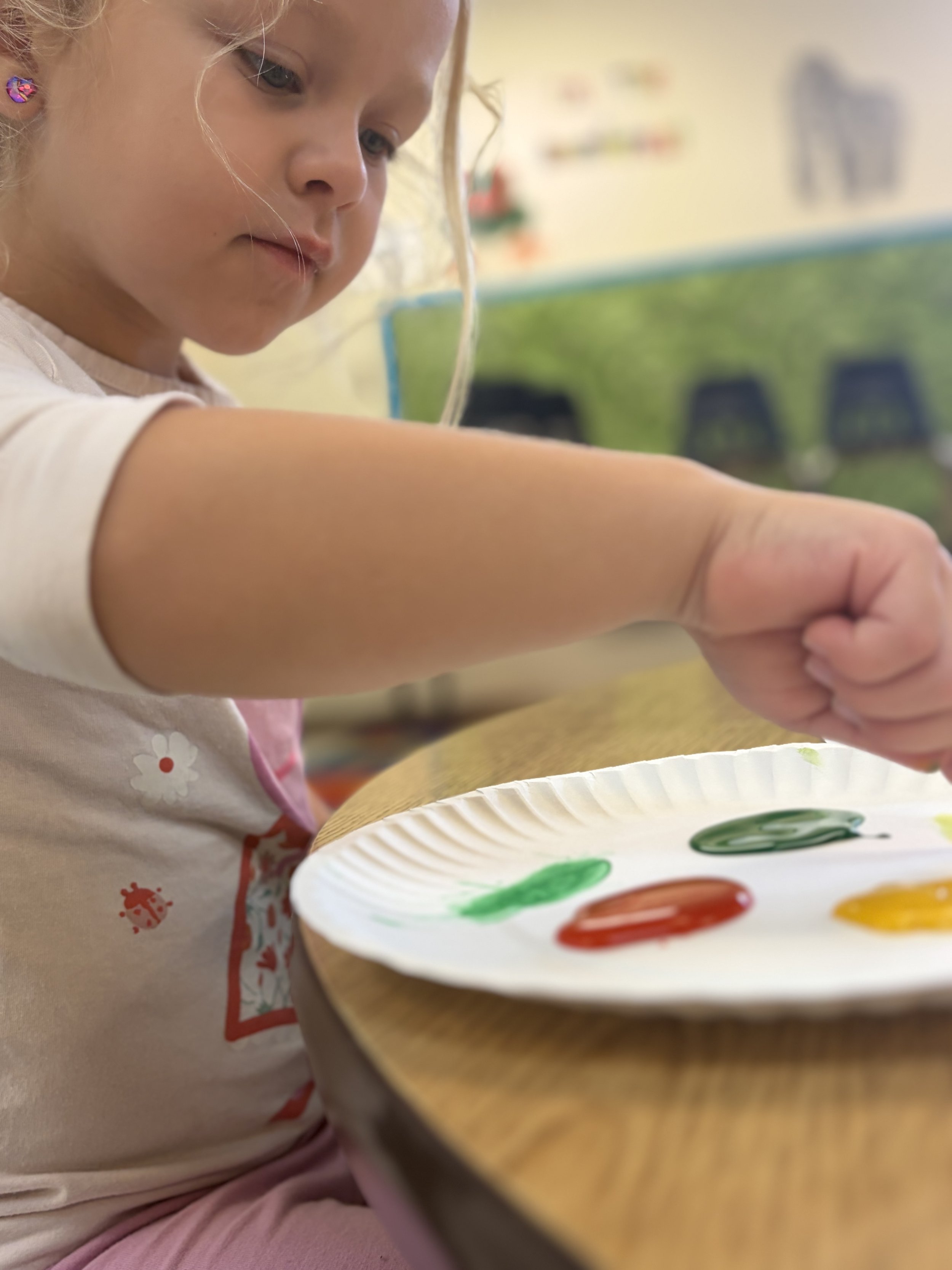 A young girl with blonde hair and butterfly-shaped earrings is painting on a paper plate with green, red, and yellow colors, sitting at a table in a classroom.