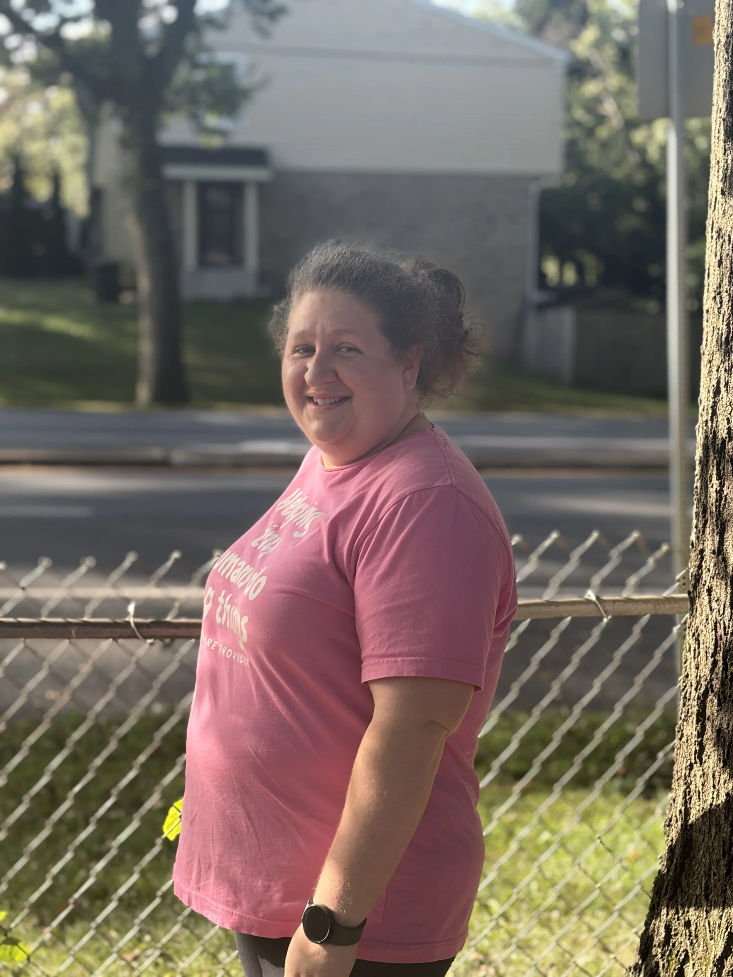 A smiling woman wearing a pink T-shirt and a black smartwatch standing outdoors near a tree and a chain-link fence, with houses and trees in the background.