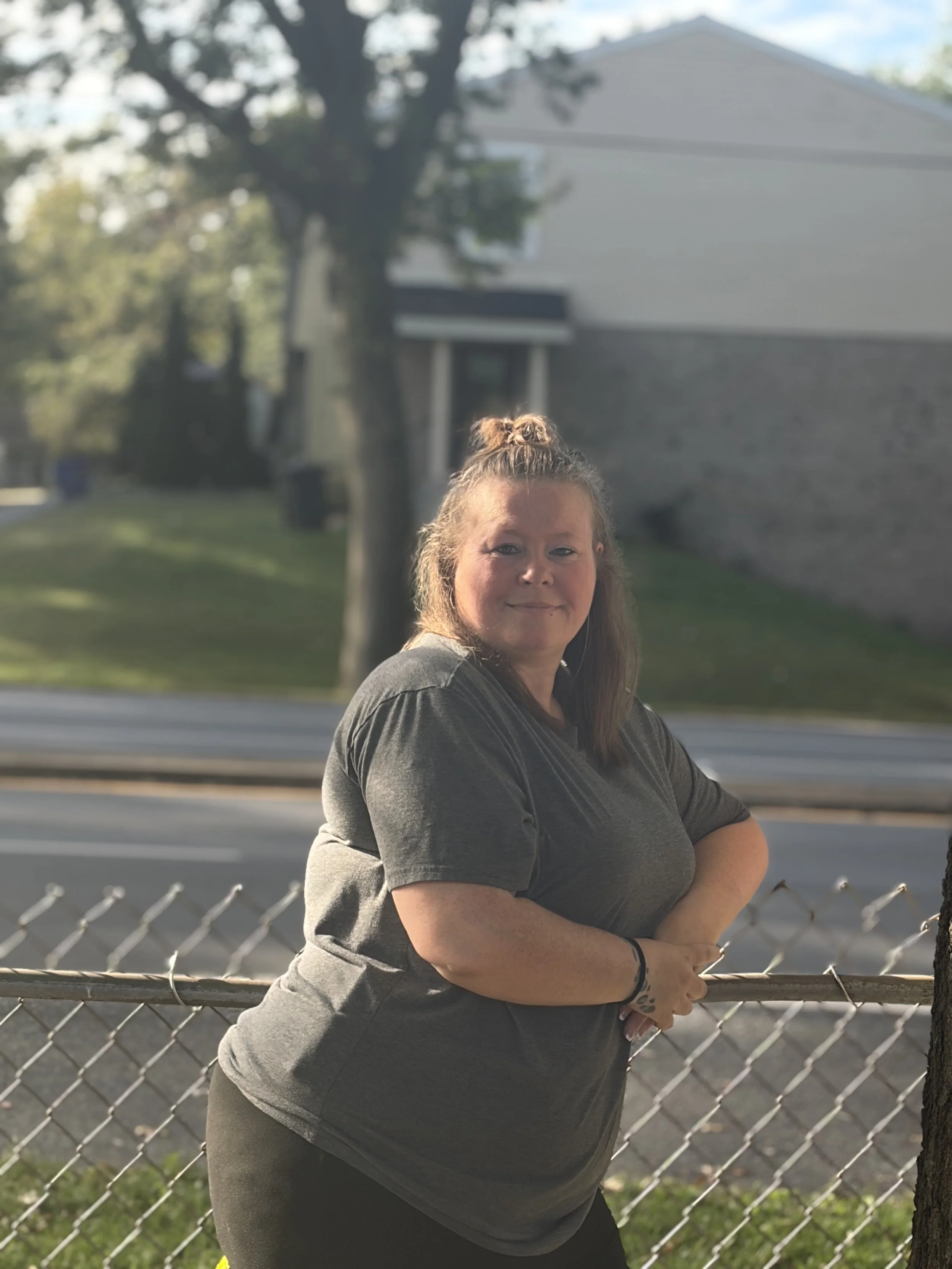 A woman with long hair tied in a bun, smiling, standing outdoors behind a chain-link fence. She is wearing a dark t-shirt, and the background includes trees, a house, and a road, with clear weather.