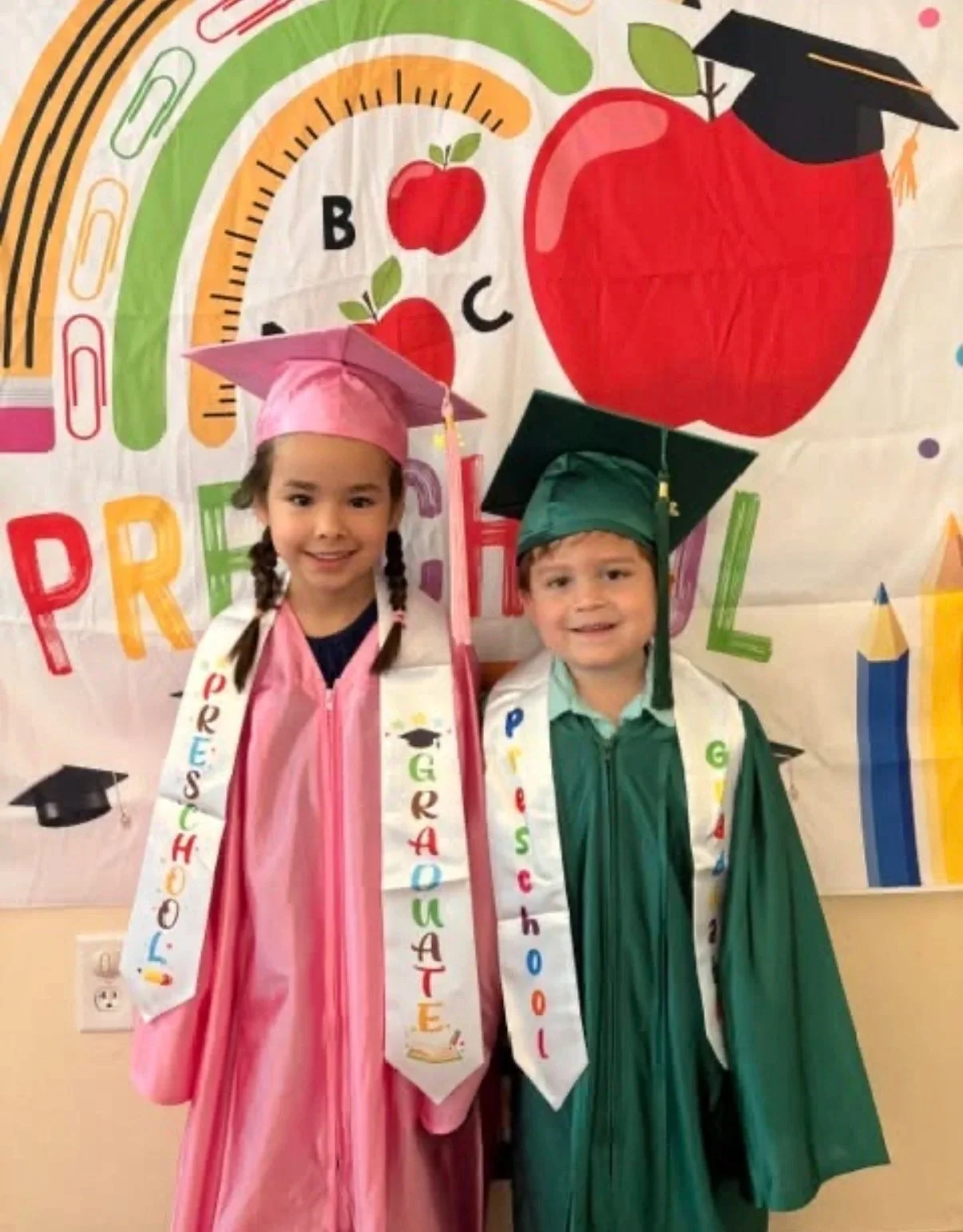 Two young children dressed in graduation caps and gowns standing in front of a colorful classroom wall decorated with apples, a rainbow, and books, celebrating a graduation event.