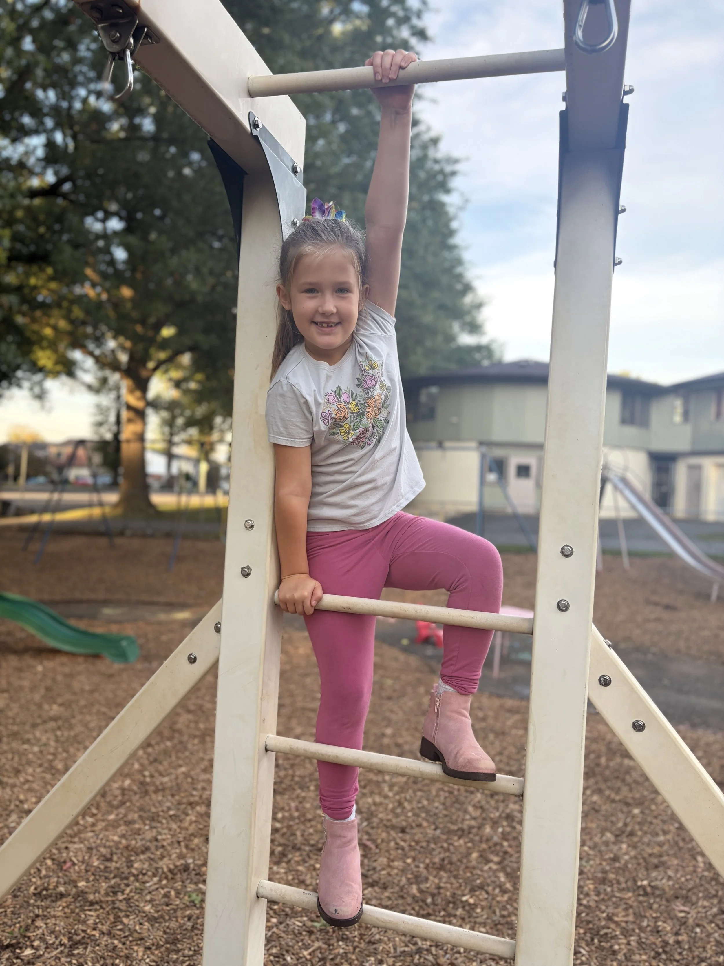 Young girl with a ponytail and floral shirt, hanging from the top of a playground slide, smiling at the camera, with trees and a building in the background.