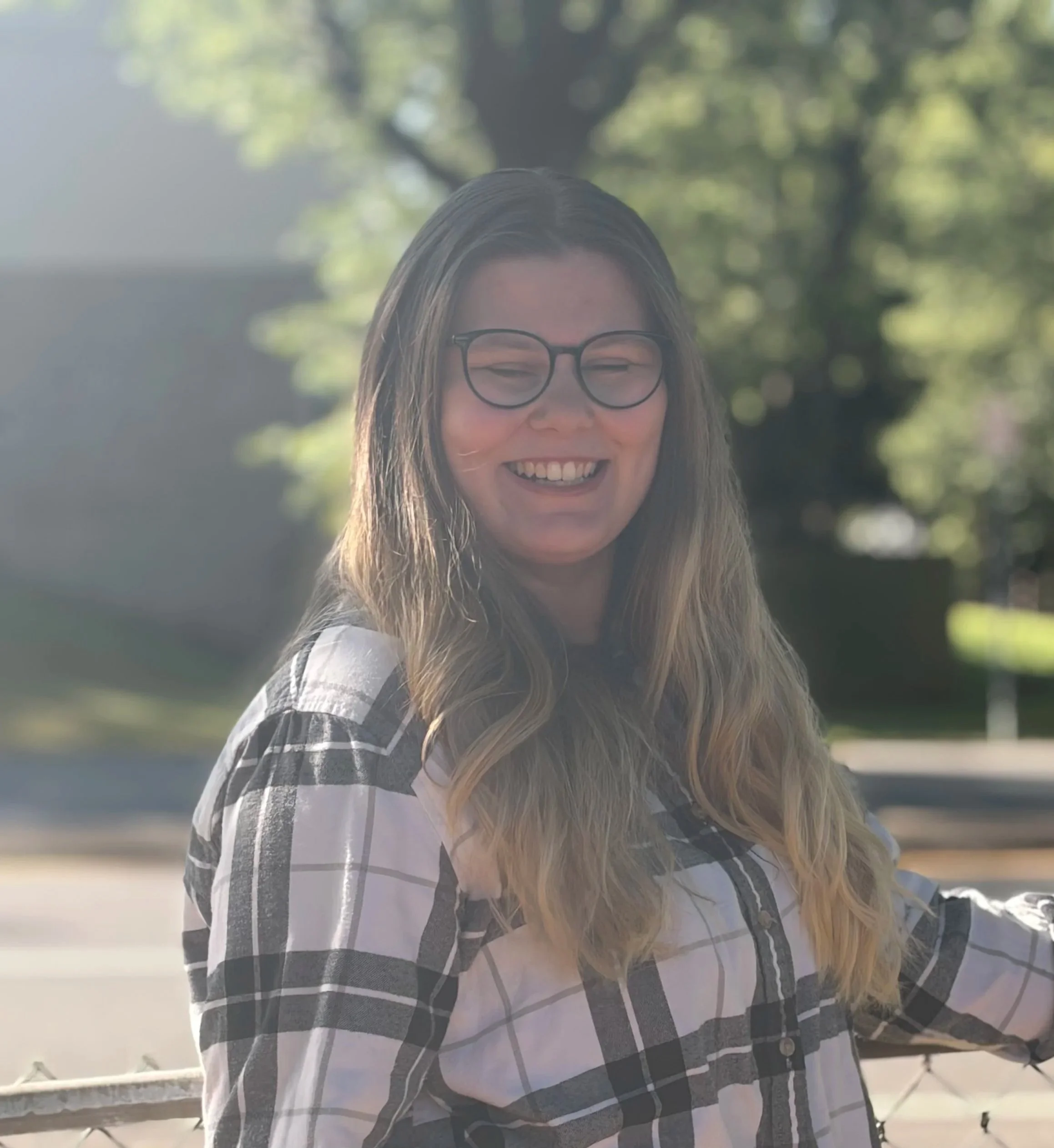 A smiling woman with long hair, glasses, and a plaid shirt standing outdoors with a blurred background of trees and sunlight.