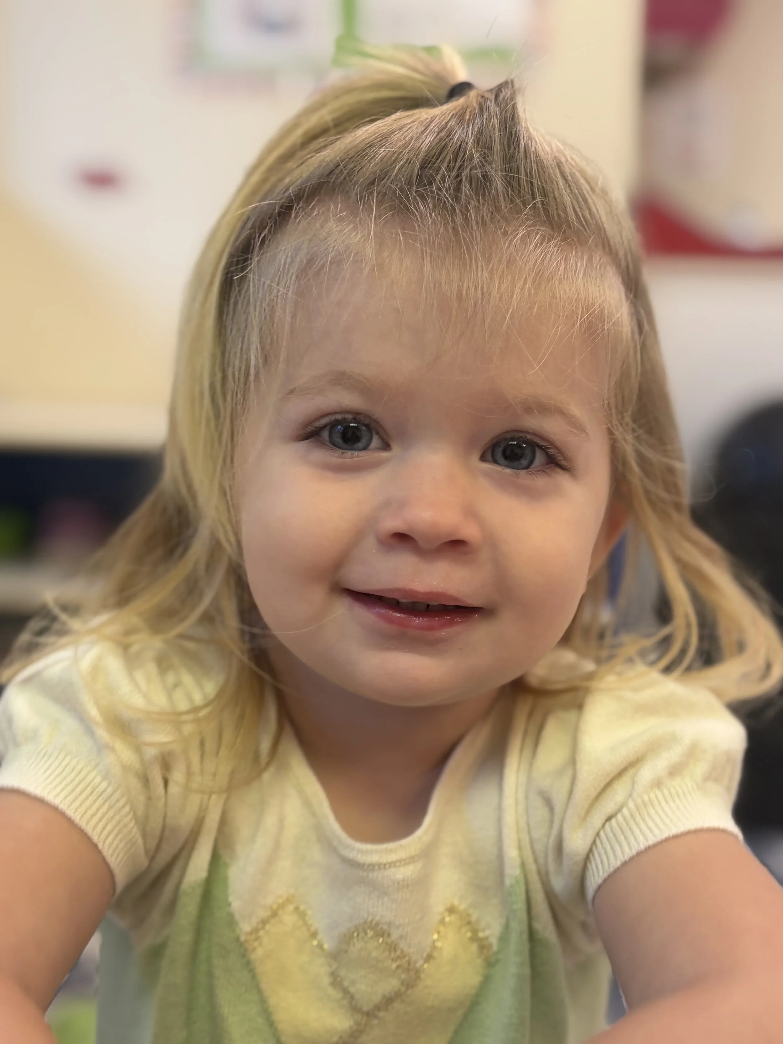 Close-up of a young girl with blonde hair, blue eyes, and a small smile, wearing a yellow shirt with a gold heart on the front. She is in an indoor setting with a blurred background.