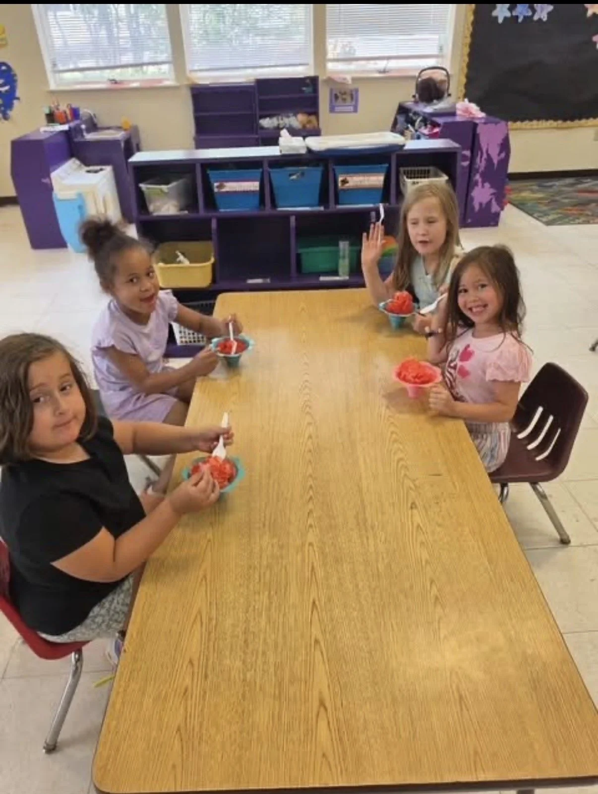Four young girls sitting at a wooden table in a classroom, eating bowls of shaved ice with red syrup. The classroom has purple shelves, large windows, and colorful decorations.