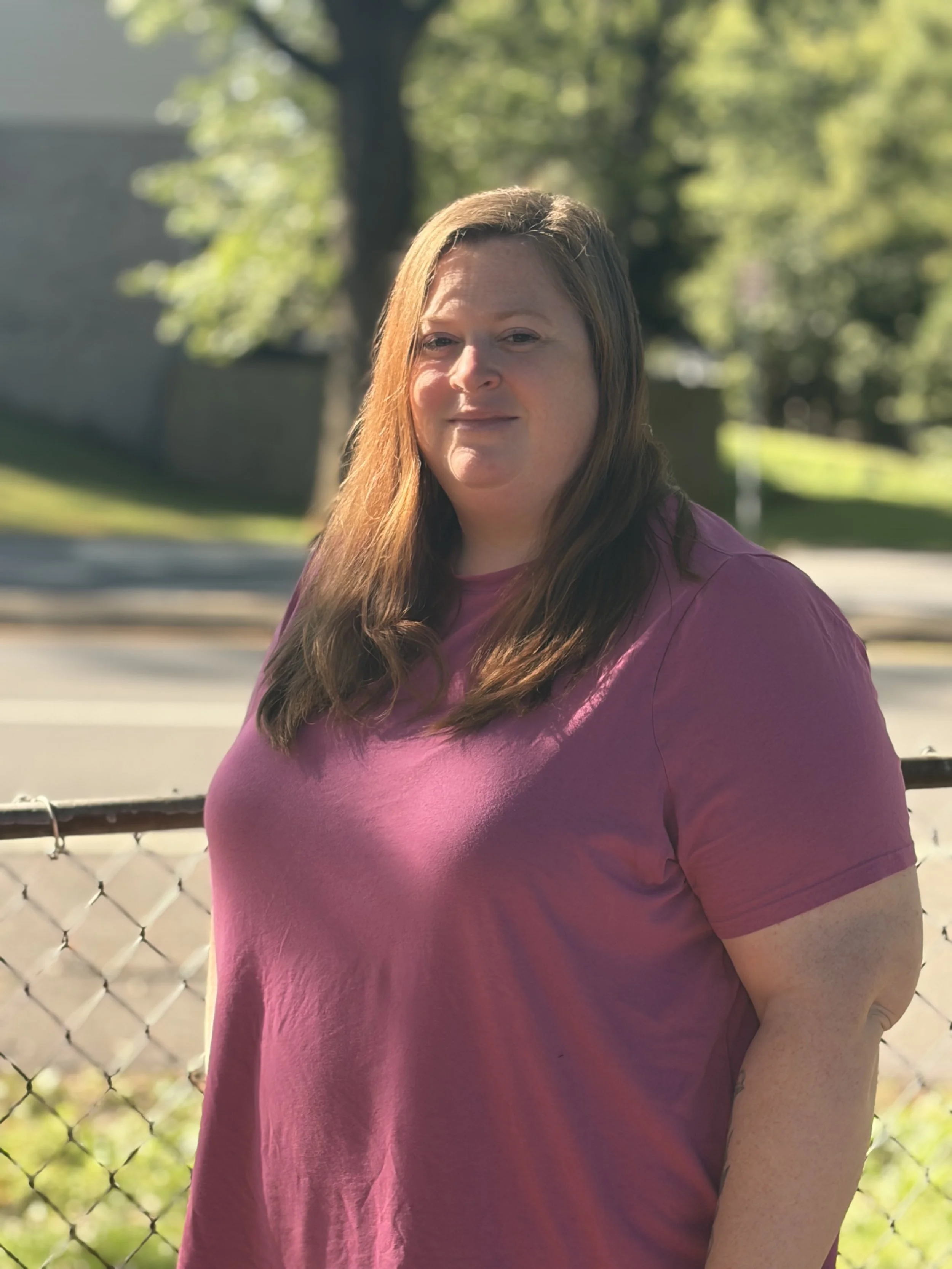 A woman with long reddish-brown hair wearing a purple shirt standing outdoors near a chain-link fence, with greenery and trees in the background.