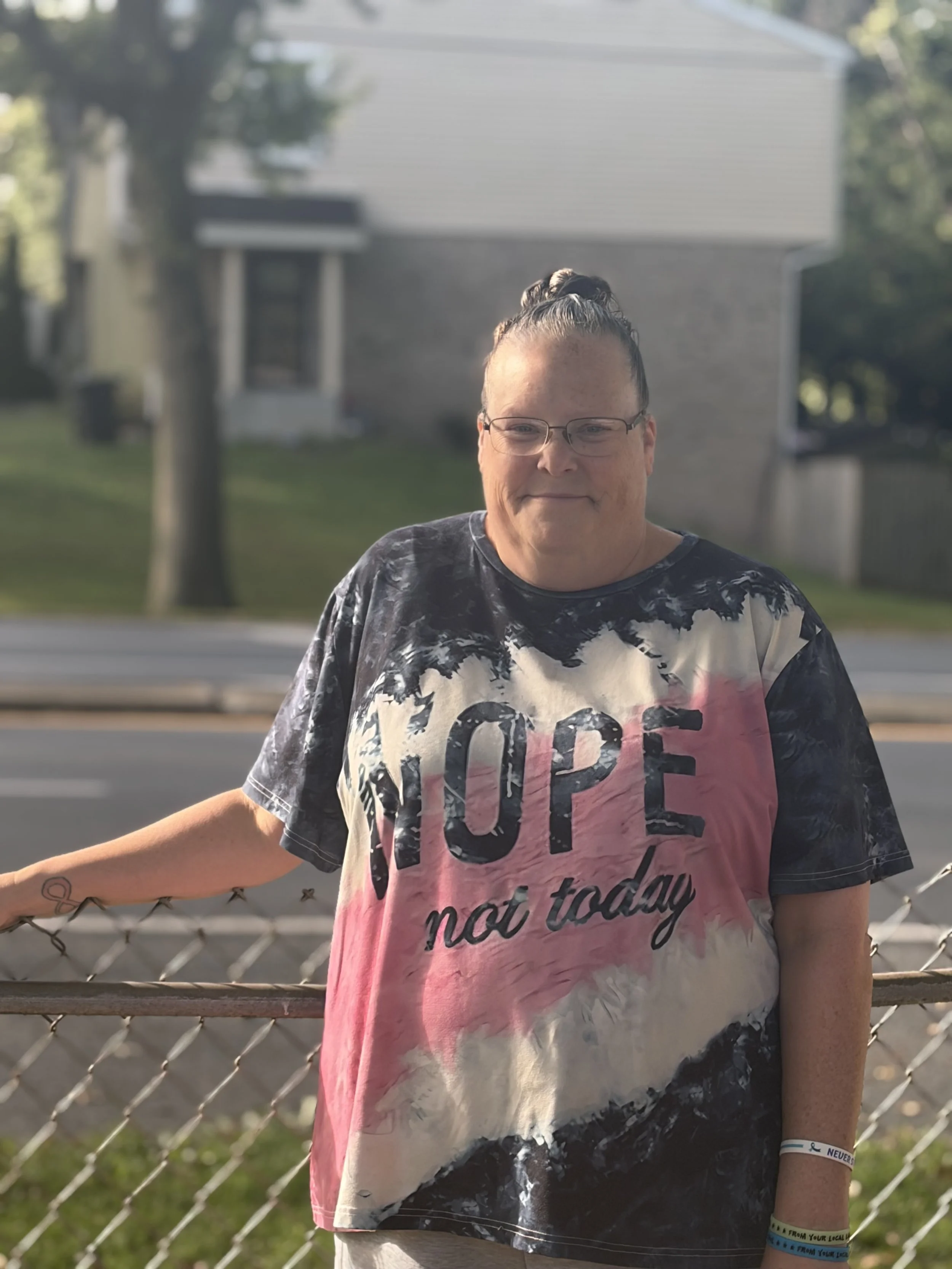 A person with glasses and braided hair standing in front of a chain-link fence outdoors, wearing a tie-dye T-shirt with the word "hope" and a wristband.
