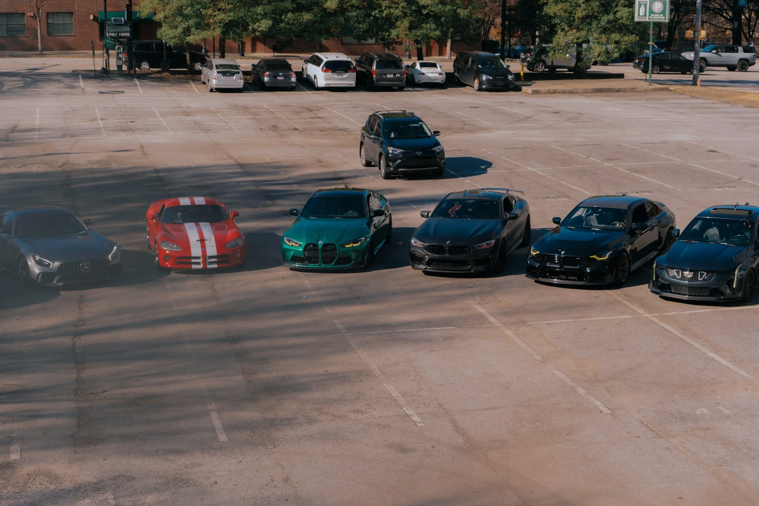 An empty parking lot with a few cars parked along the back row and some cars parked close together in the front. The parking lot is adjacent to a building with trees in the background.