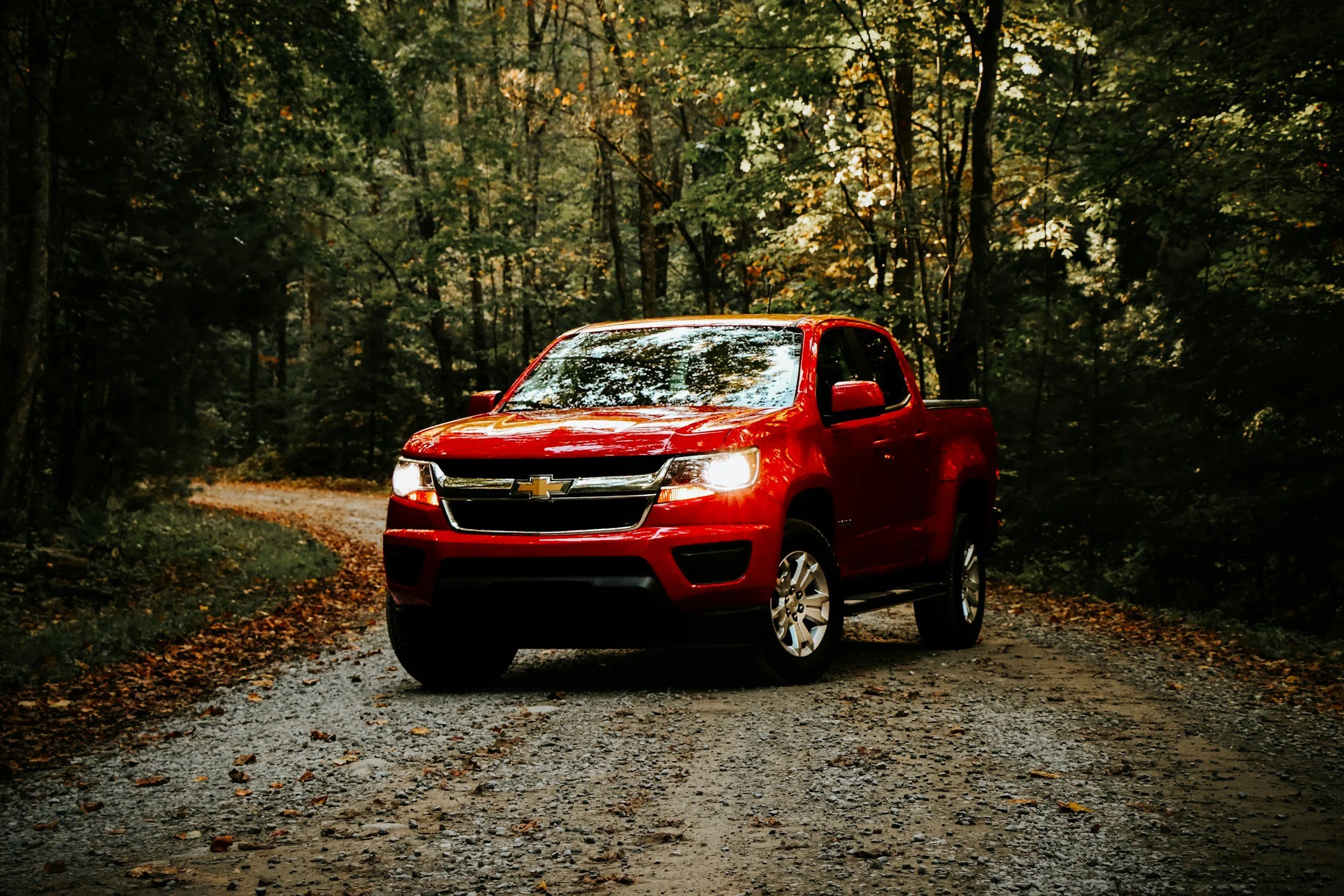 Red Chevrolet pickup truck driving on a gravel forest path surrounded by green trees with autumn leaves.
