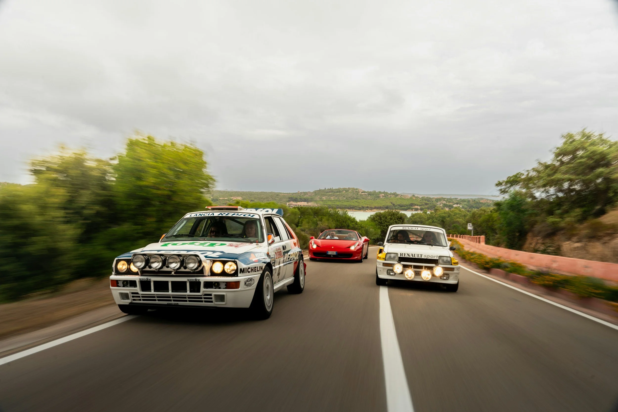 Multiple rally cars racing on a winding scenic road with greenery on both sides and cloudy sky overhead.
