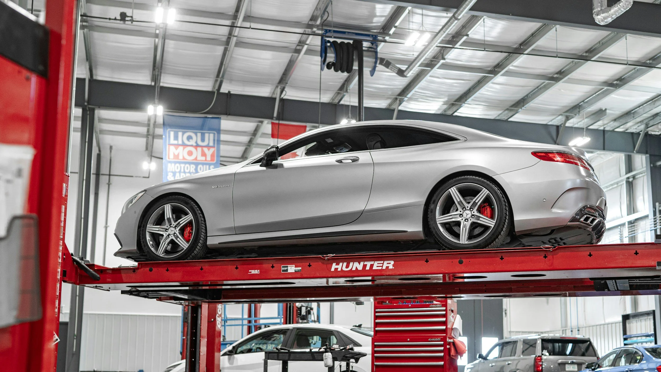 A silver Mercedes-Benz sports car on a red hydraulic lift inside an automotive service center. In the background, there are tools, hanging equipment, and a sign for Lubri Moly motor oils.