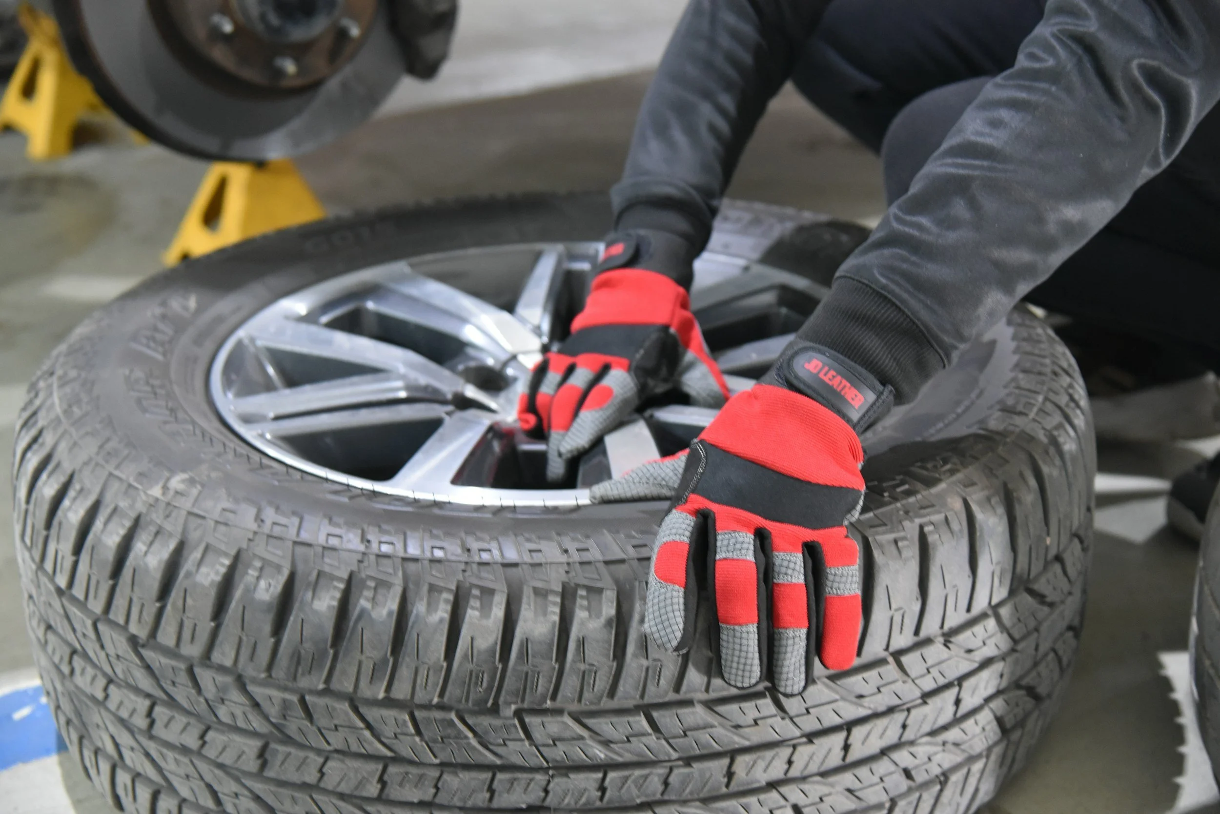 Person wearing black jacket, red and black gloves, working on a car tire in a garage. The person is placing or removing a silver rim from the tire, with a yellow jack and workshop setting in the background.