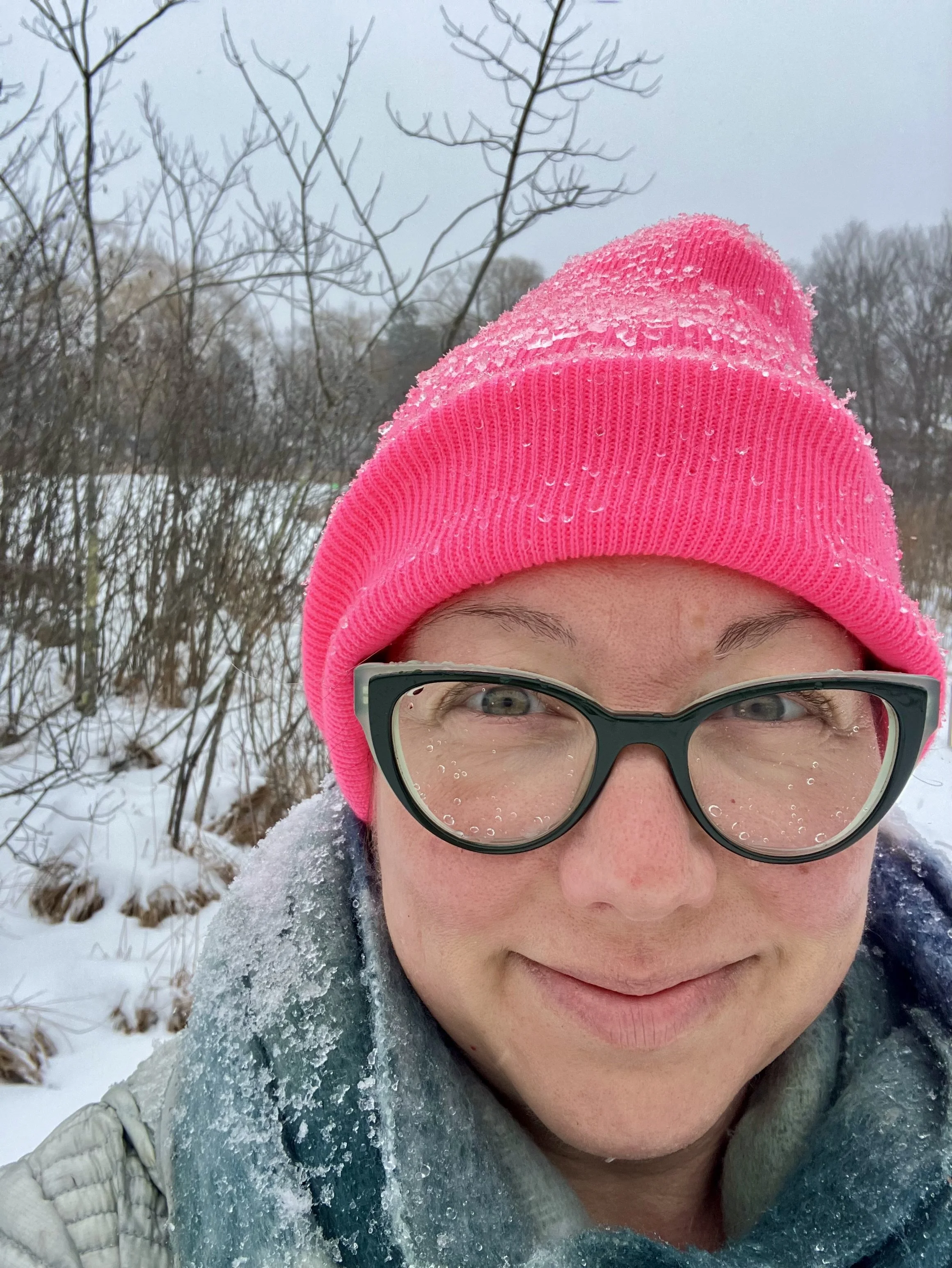 Close-up of a person wearing a bright pink knit cap with frost and snowflakes on it, glasses with water droplets, and winter clothing in a snowy outdoor setting with leafless trees in the background.