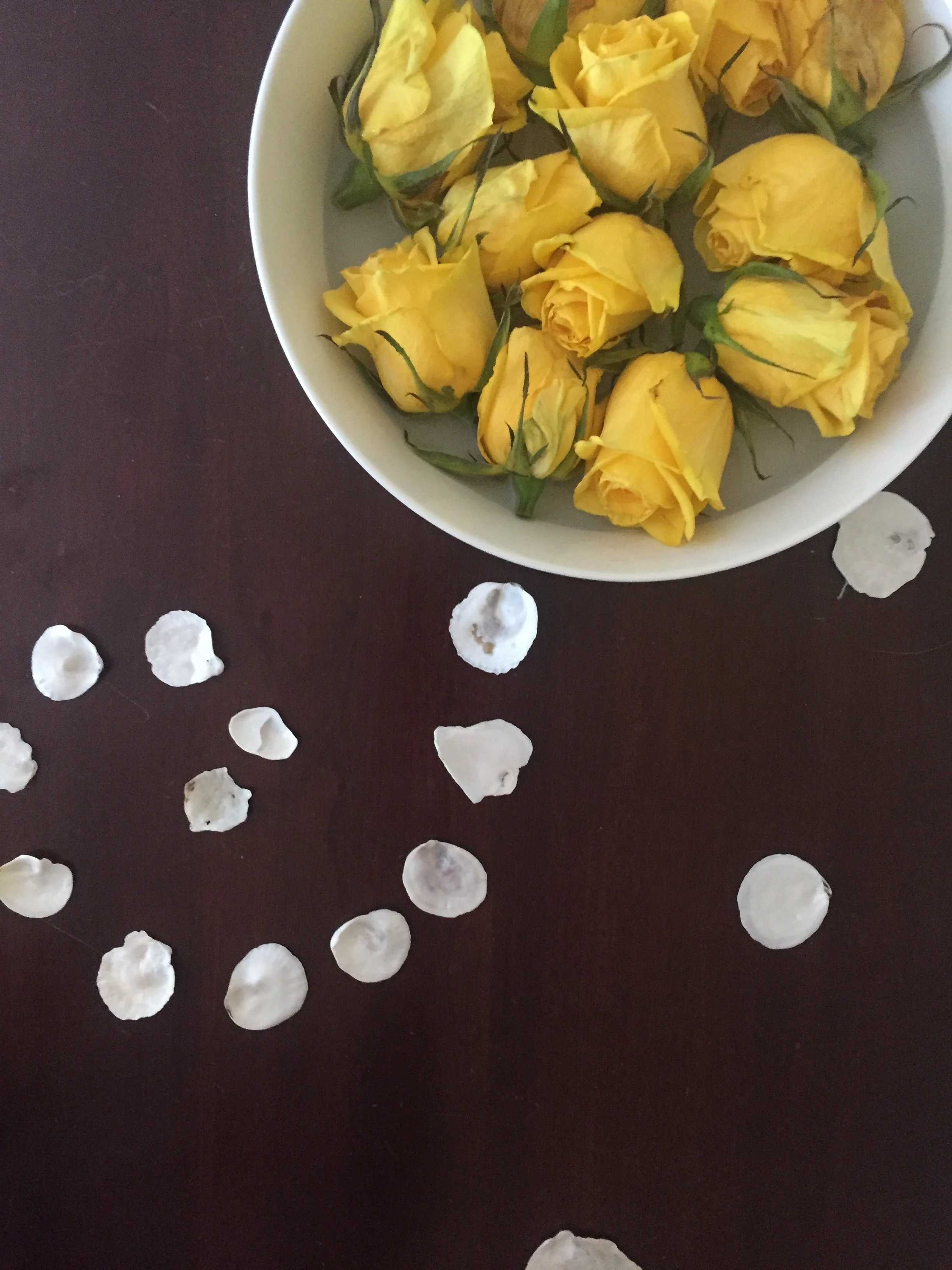 A white bowl filled with yellow rose petals, placed on a dark wooden surface with white flower petals scattered around.