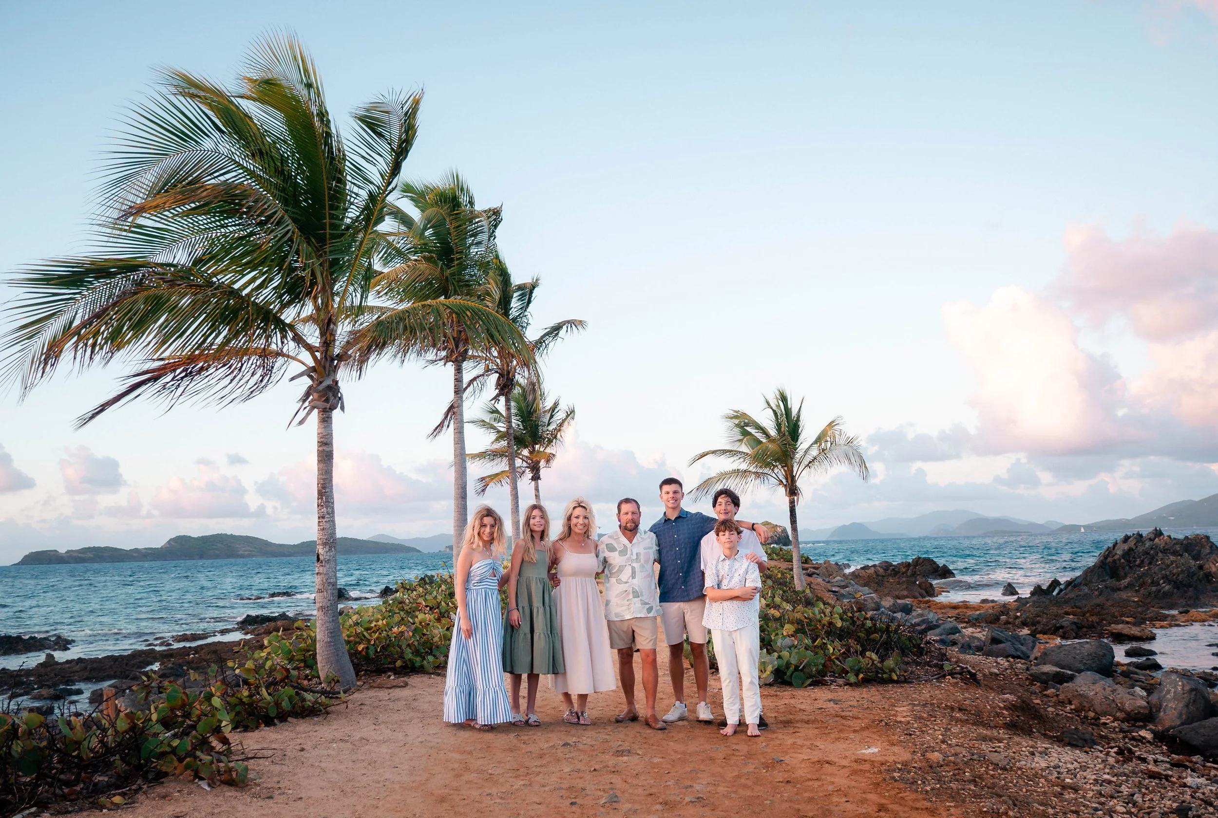 A group of nine people standing on a rocky beach with palm trees, ocean, and distant mountains in the background during sunset.