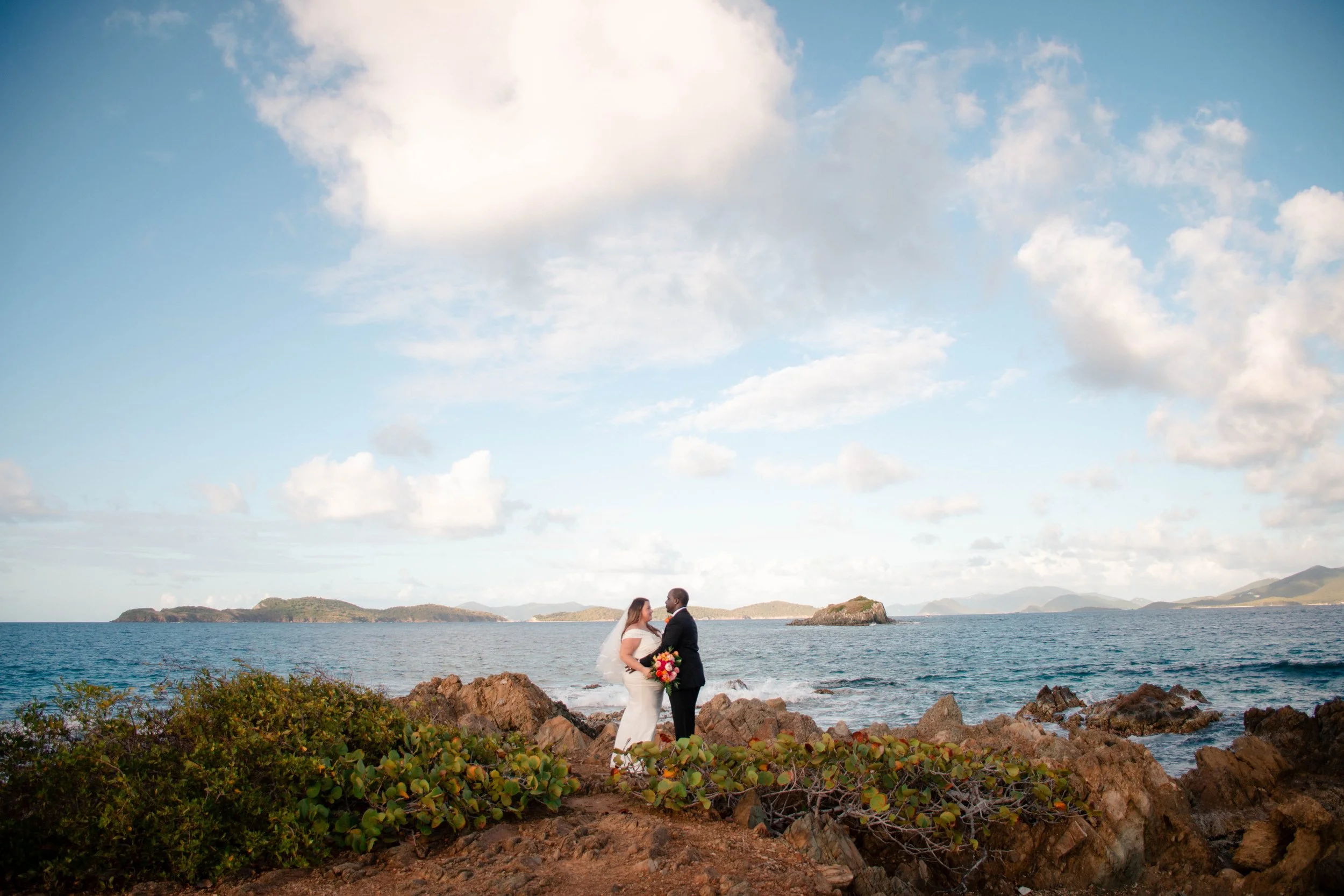 A bride and groom standing on rocks near the ocean, holding hands and facing each other, with small islands and a partly cloudy sky in the background.