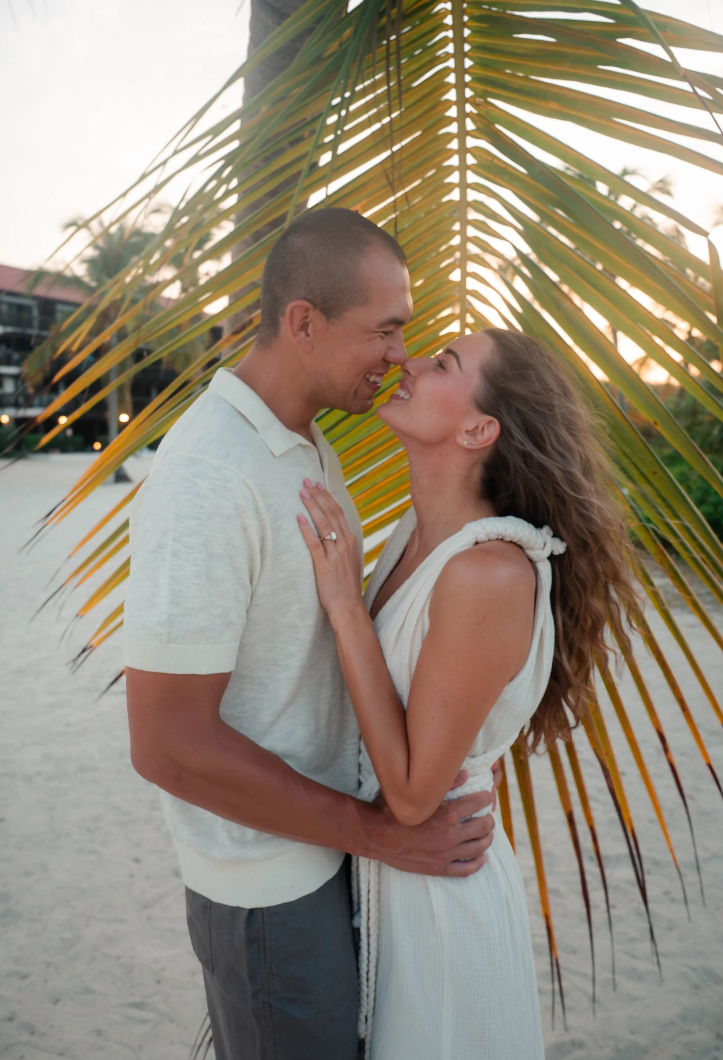 A couple standing close on a beach with palm leaves behind them, about to kiss, during sunset.