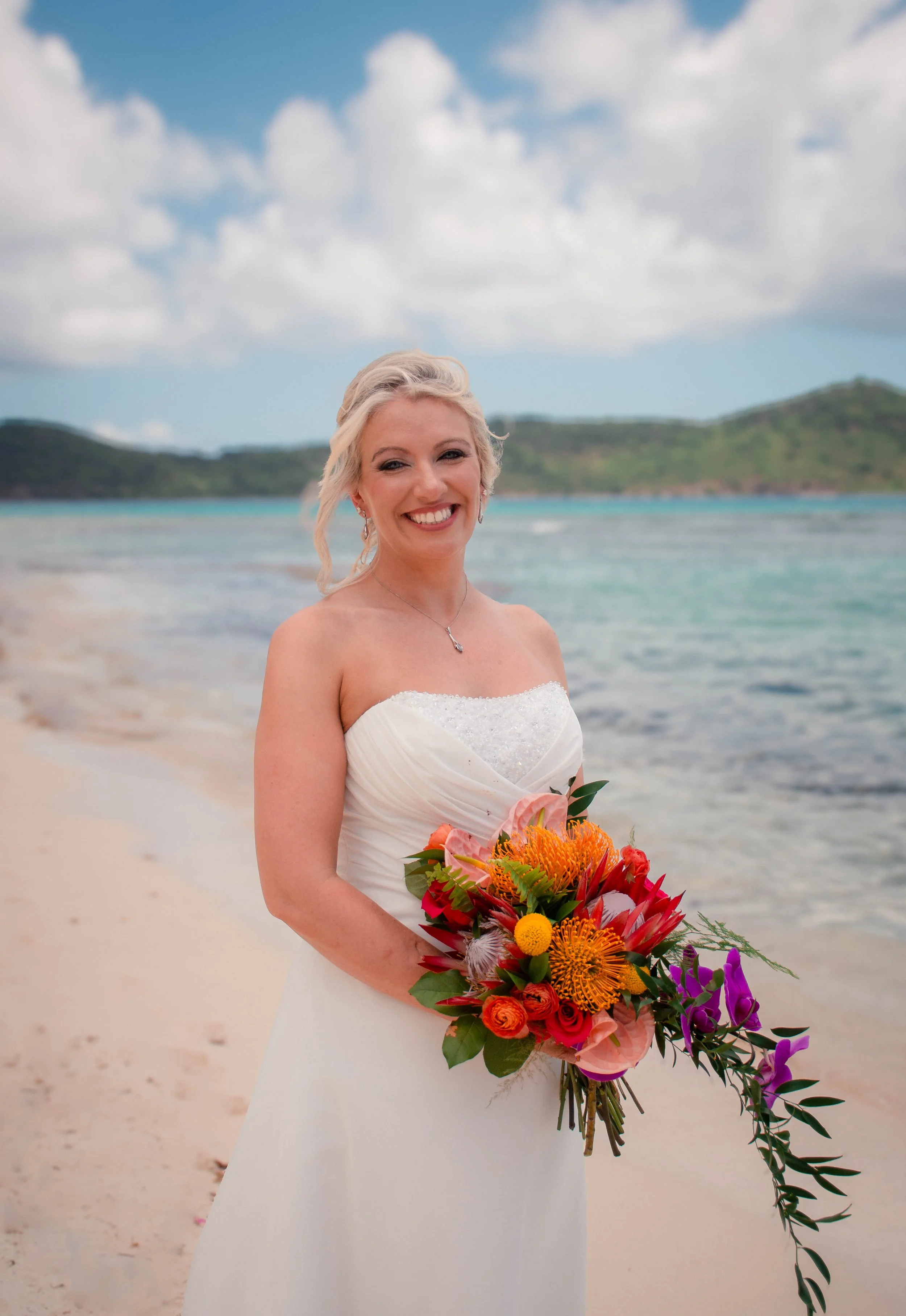 A woman in a wedding dress holding a colorful bouquet of flowers on the beach with the ocean, hills, and blue sky with clouds in the background.