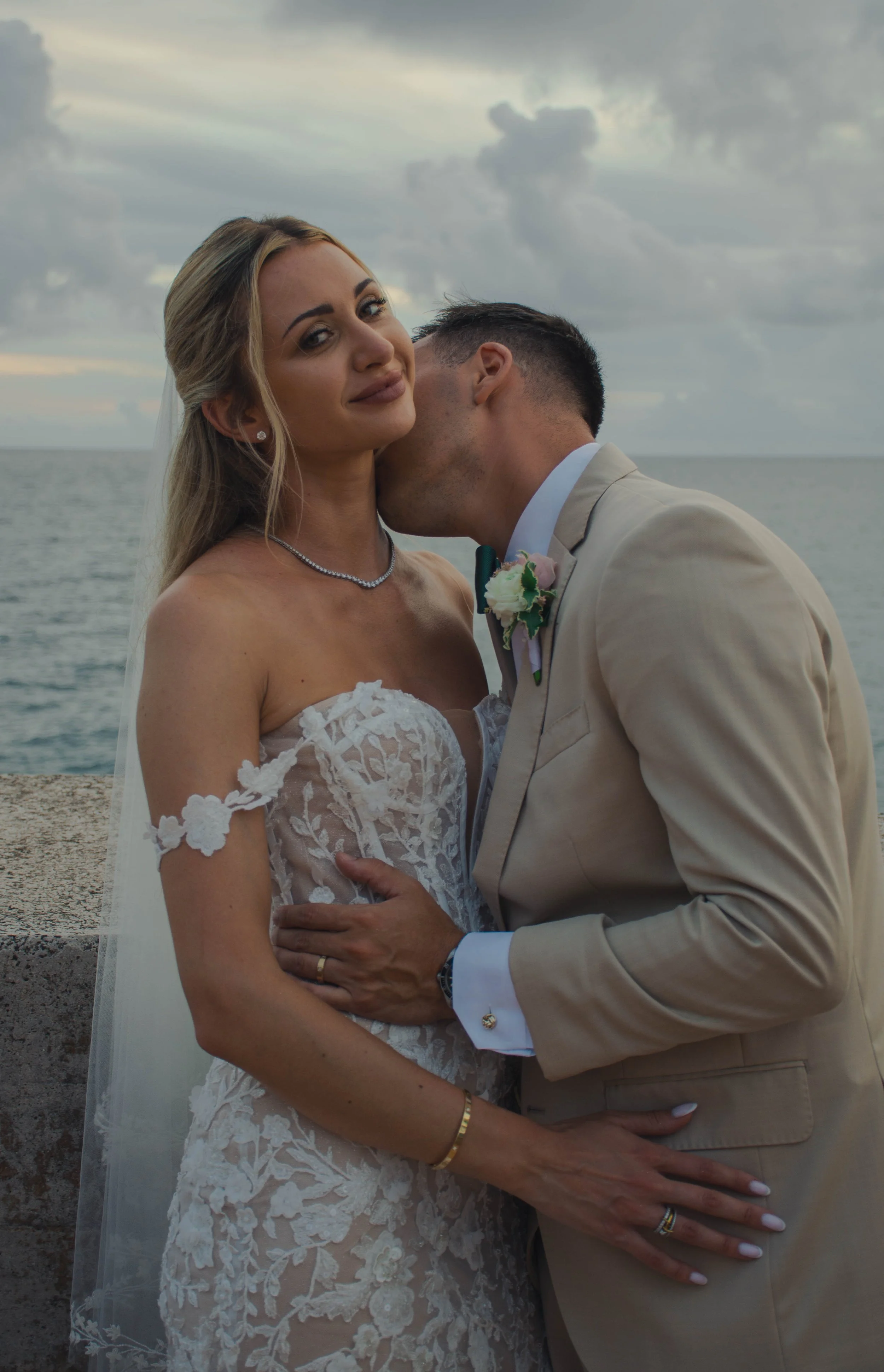 A newlywed couple standing by the ocean, with the groom kissing the bride's cheek. She is wearing a lace wedding dress and jewelry, while he is dressed in a beige suit with a boutonniere. The sky is cloudy.