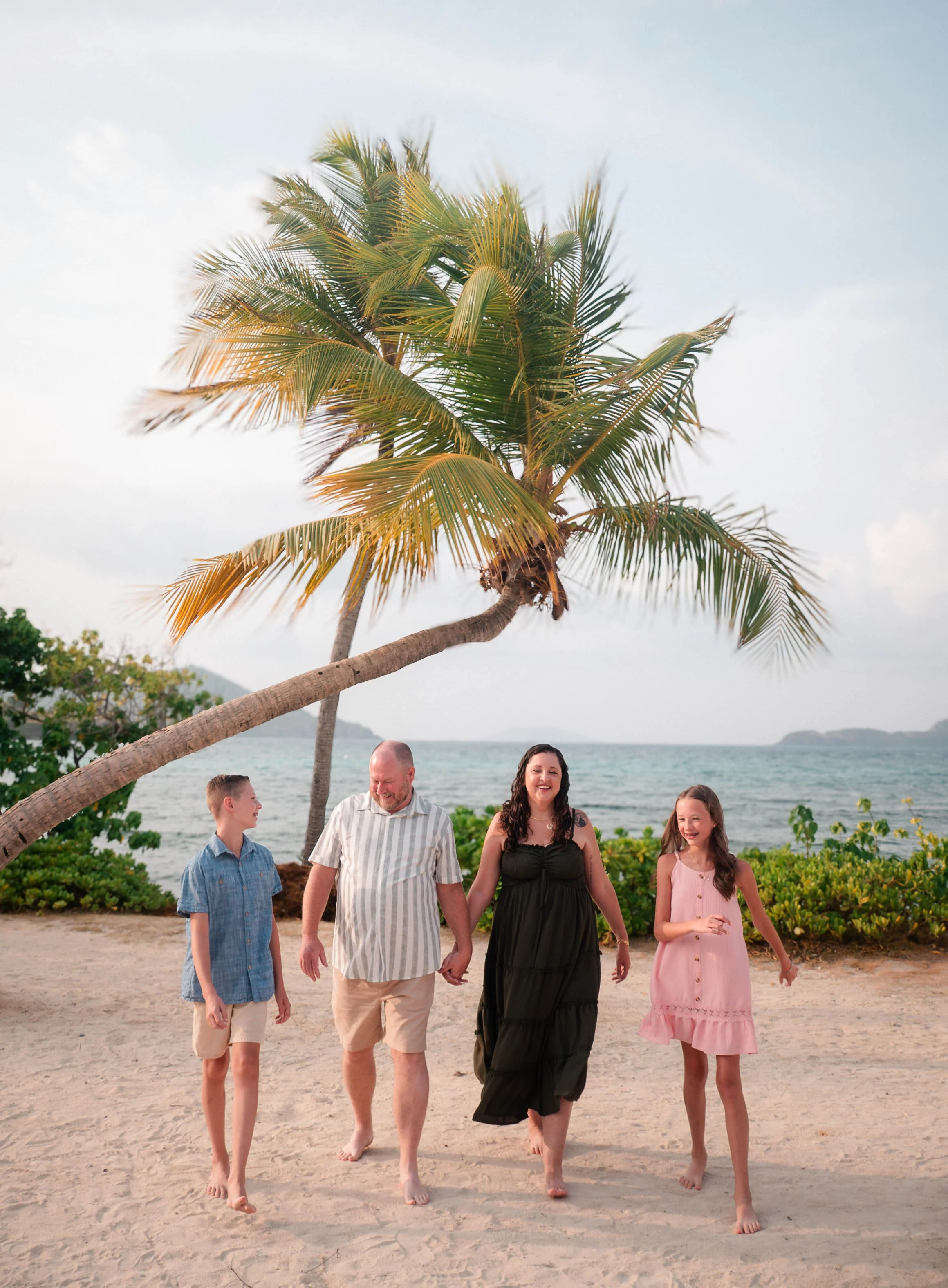 A family of five walking on the beach holding hands, with the ocean and a hillside in the background, under a clear sky and a large palm tree.