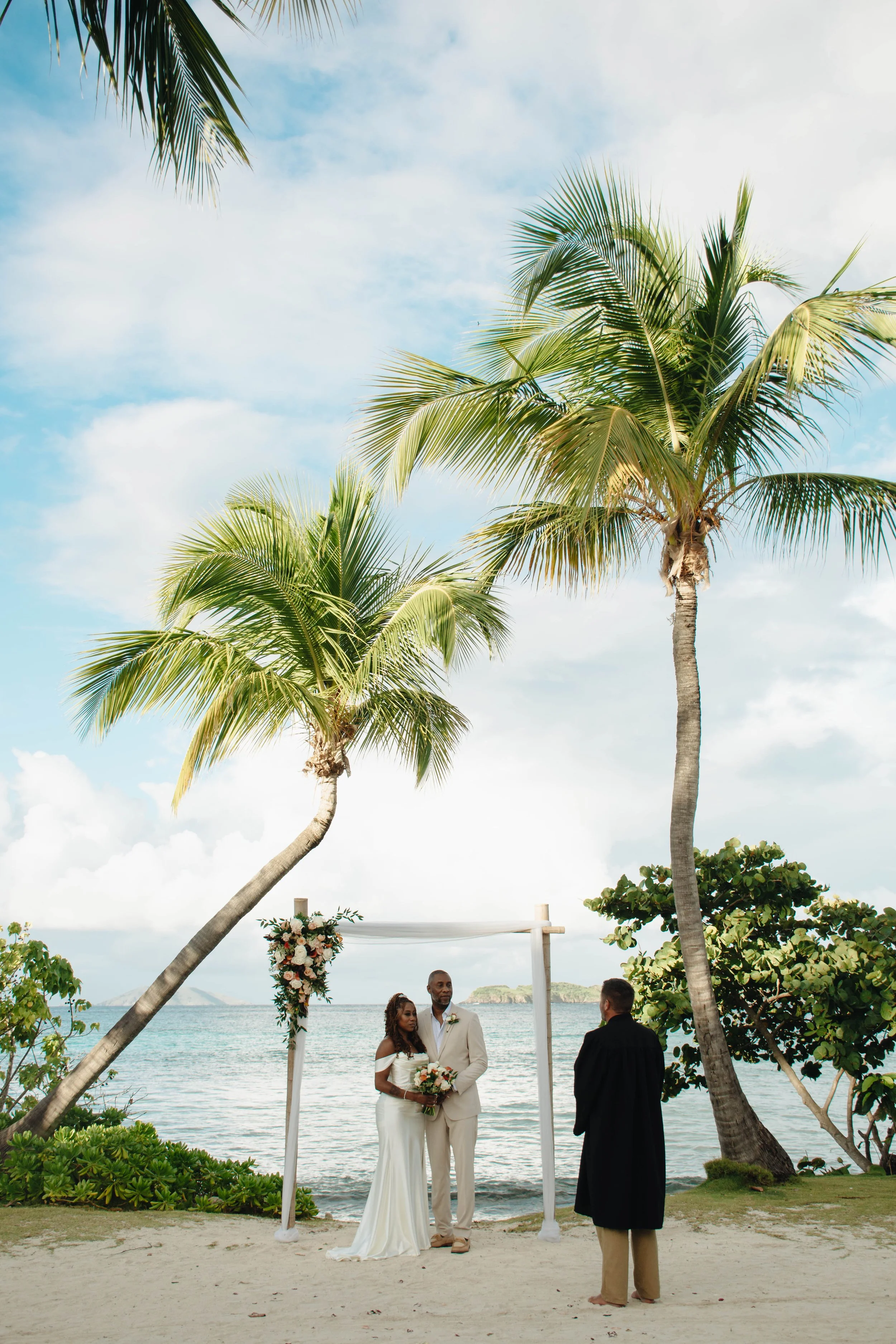 A wedding ceremony on the beach with two palm trees framing a couple exchanging vows. The bride is holding a bouquet, and the officiant stands to the right, wearing traditional robes. The backdrop includes the ocean and a distant island under a partl