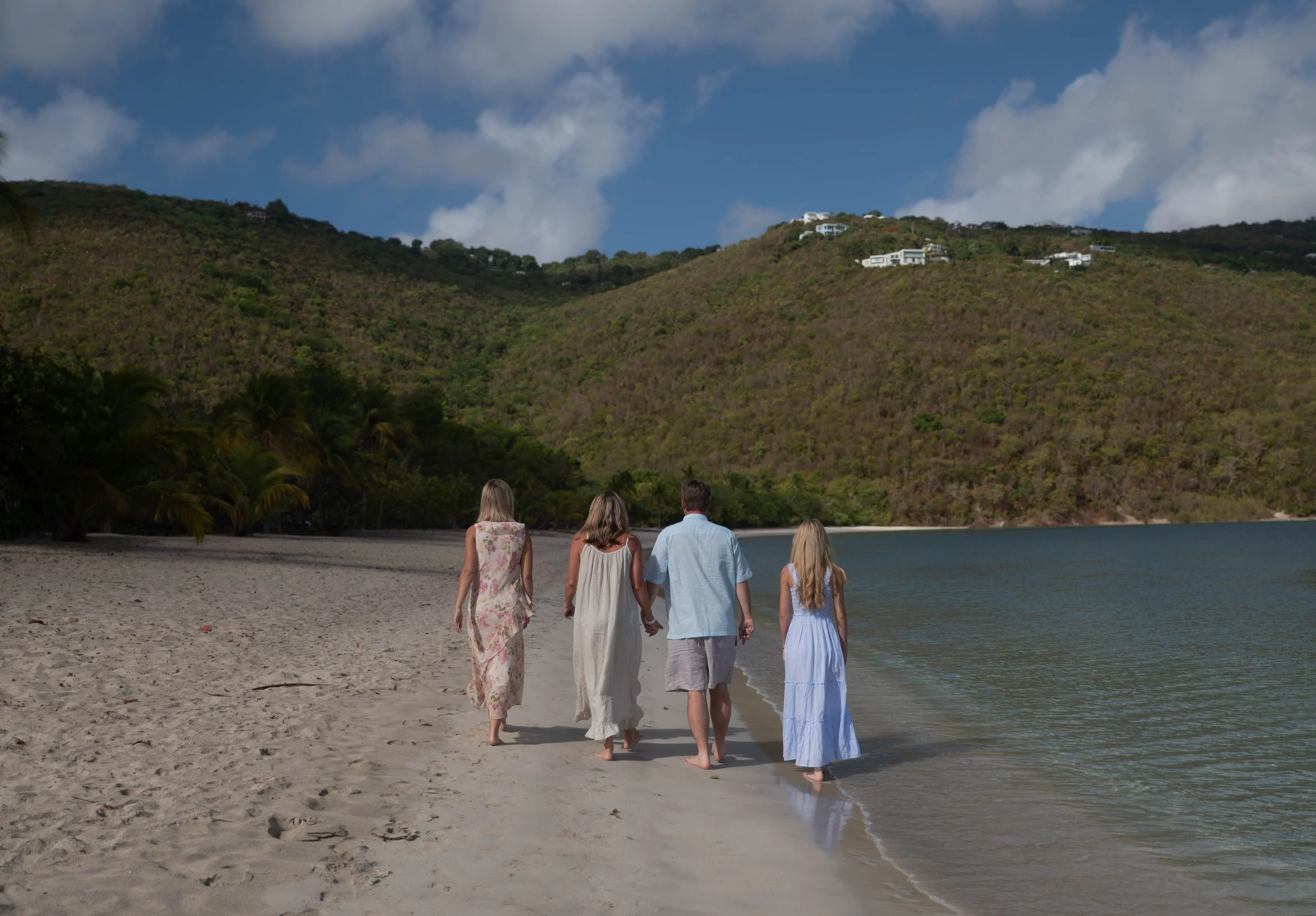 A group of five people walking along a beach by a lake or ocean, with a green hillside in the background and modern white houses on the hill. The sky is partly cloudy.