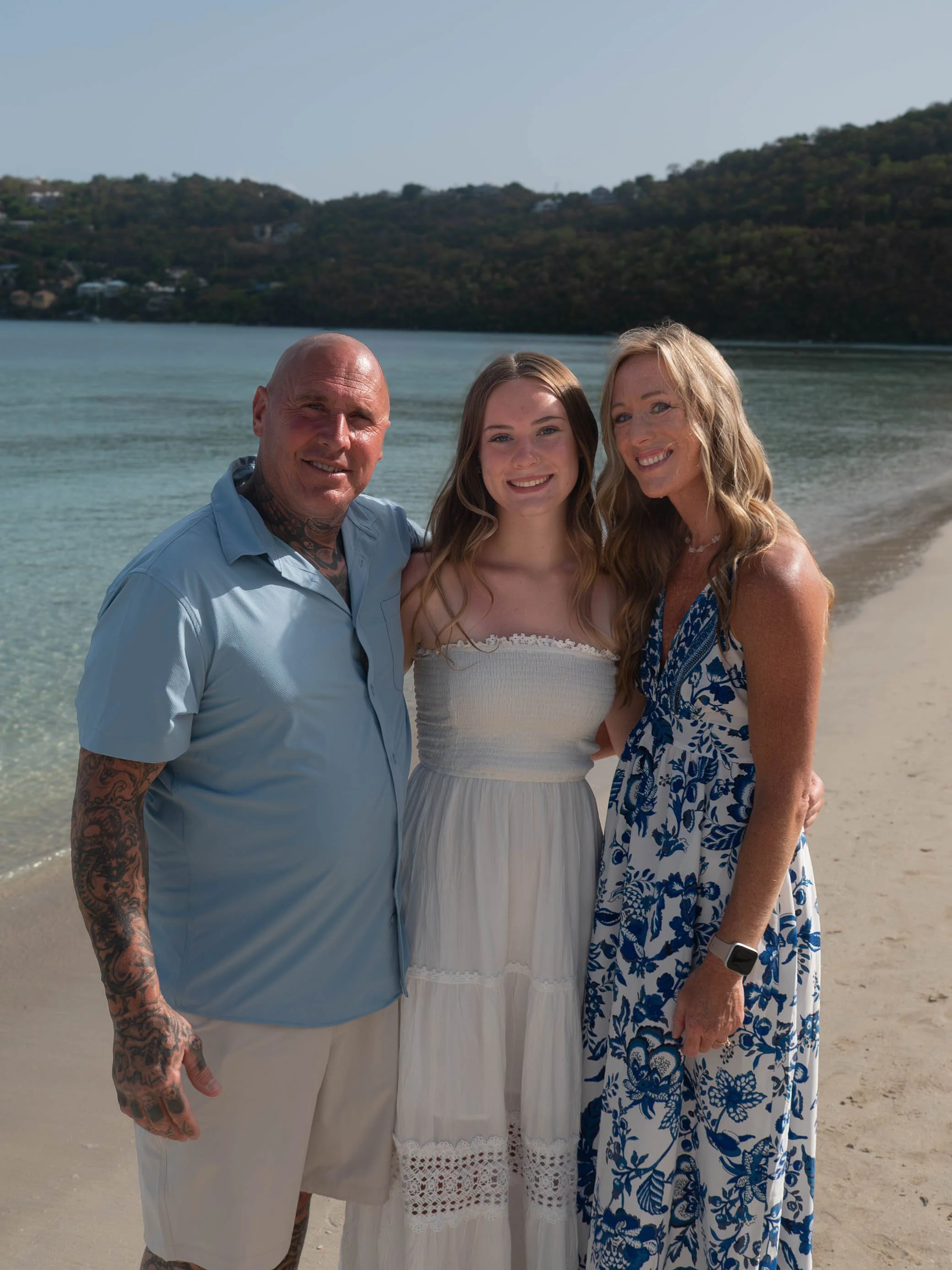A family of three standing on a beach with water and hills in the background. The man has tattoos on his arm and is wearing a light blue shirt. The young woman is wearing a white dress, and the woman is wearing a blue and white patterned dress.