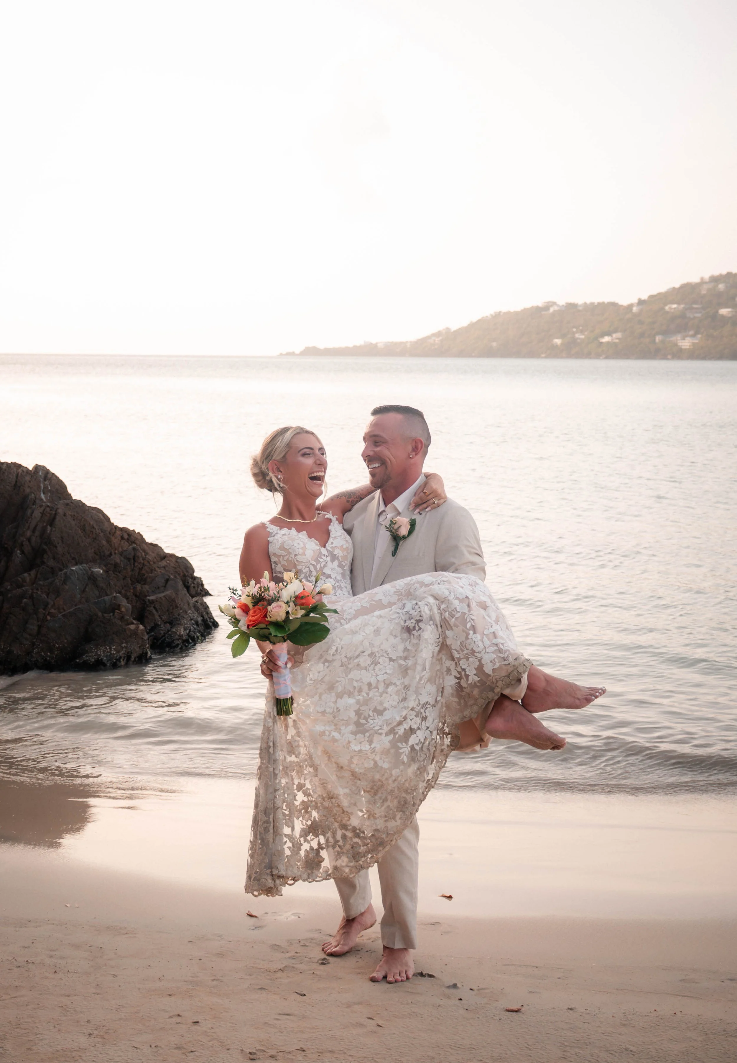 A newlywed couple at the beach, with the man lifting the woman, both smiling and laughing, holding a bouquet of flowers, with water, rocks, and a hill in the background during sunset.