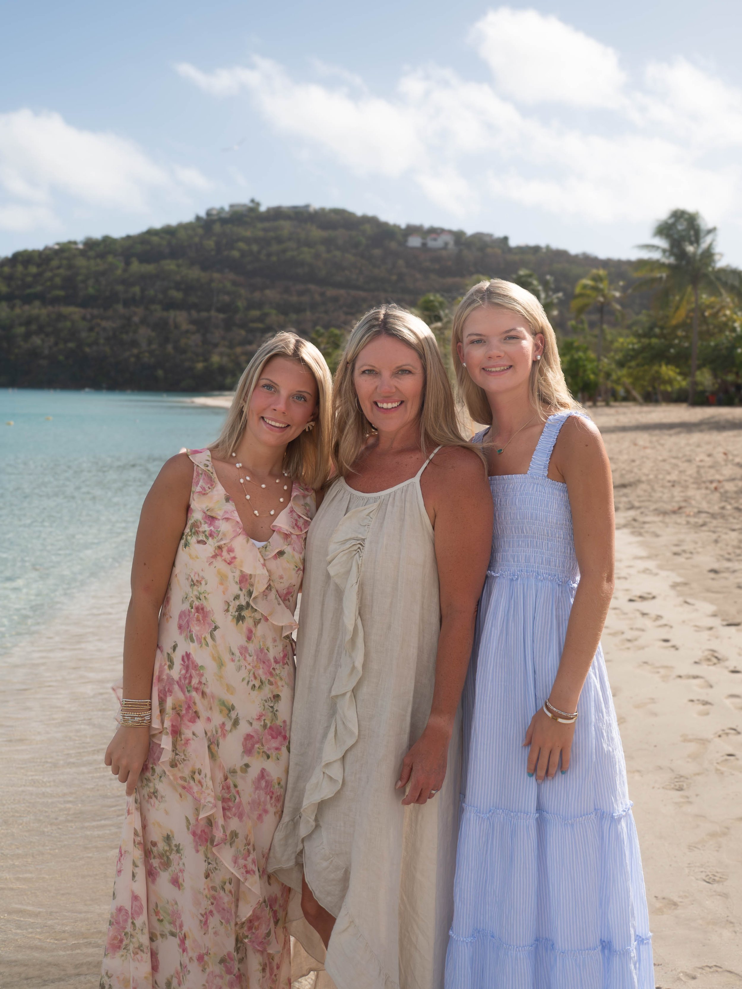 Three women standing together on a beach with a hill and trees in the background. All are smiling and dressed in light, summery dresses.