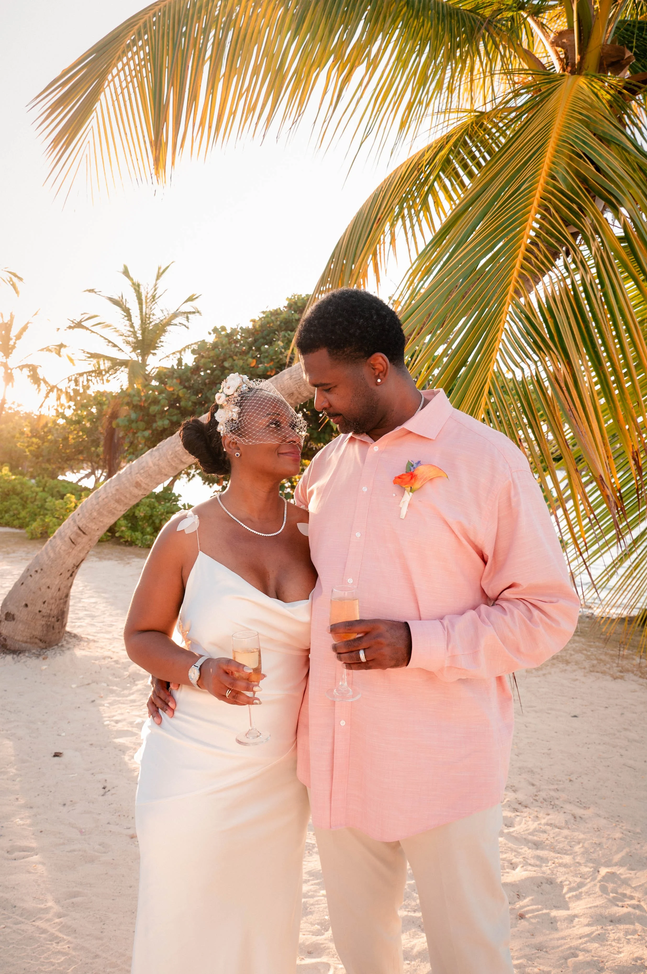 A smiling woman and man standing close together on a beach, holding glasses of champagne, under palm trees with sunset behind them, during a celebration or wedding.