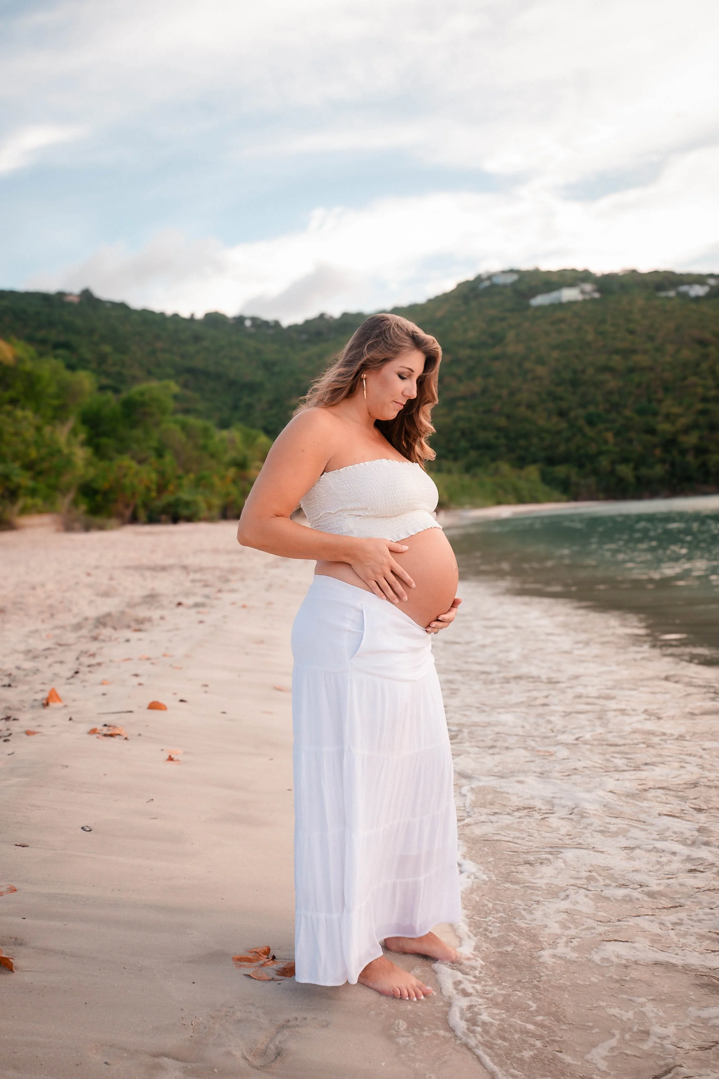 A pregnant woman in a white strapless top and long white skirt standing barefoot on a sandy beach near the water, with green hills and a cloudy sky in the background.