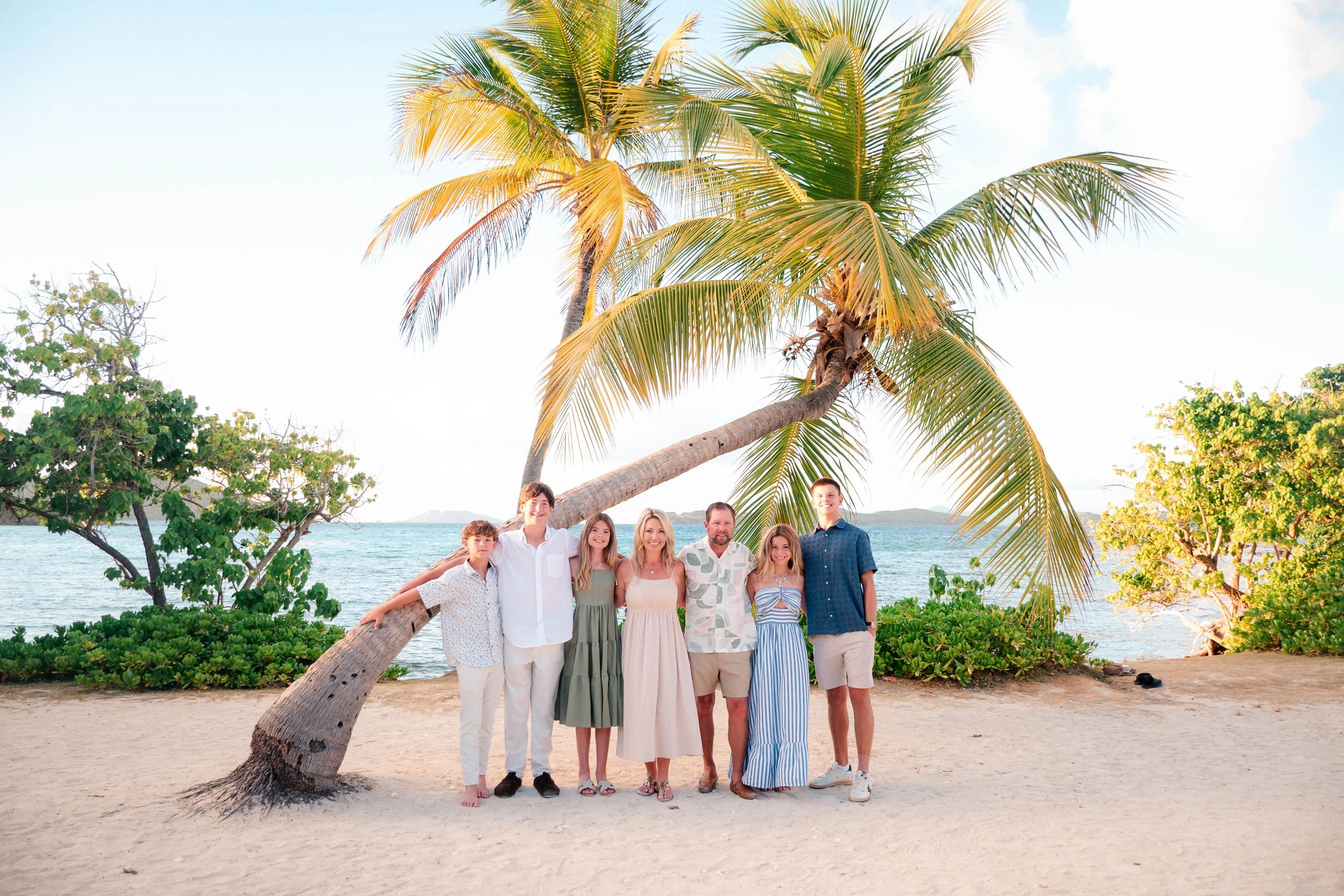 A group of nine people standing together on a beach with a large palm tree leaning over them, water and mountains in the background, during daytime.