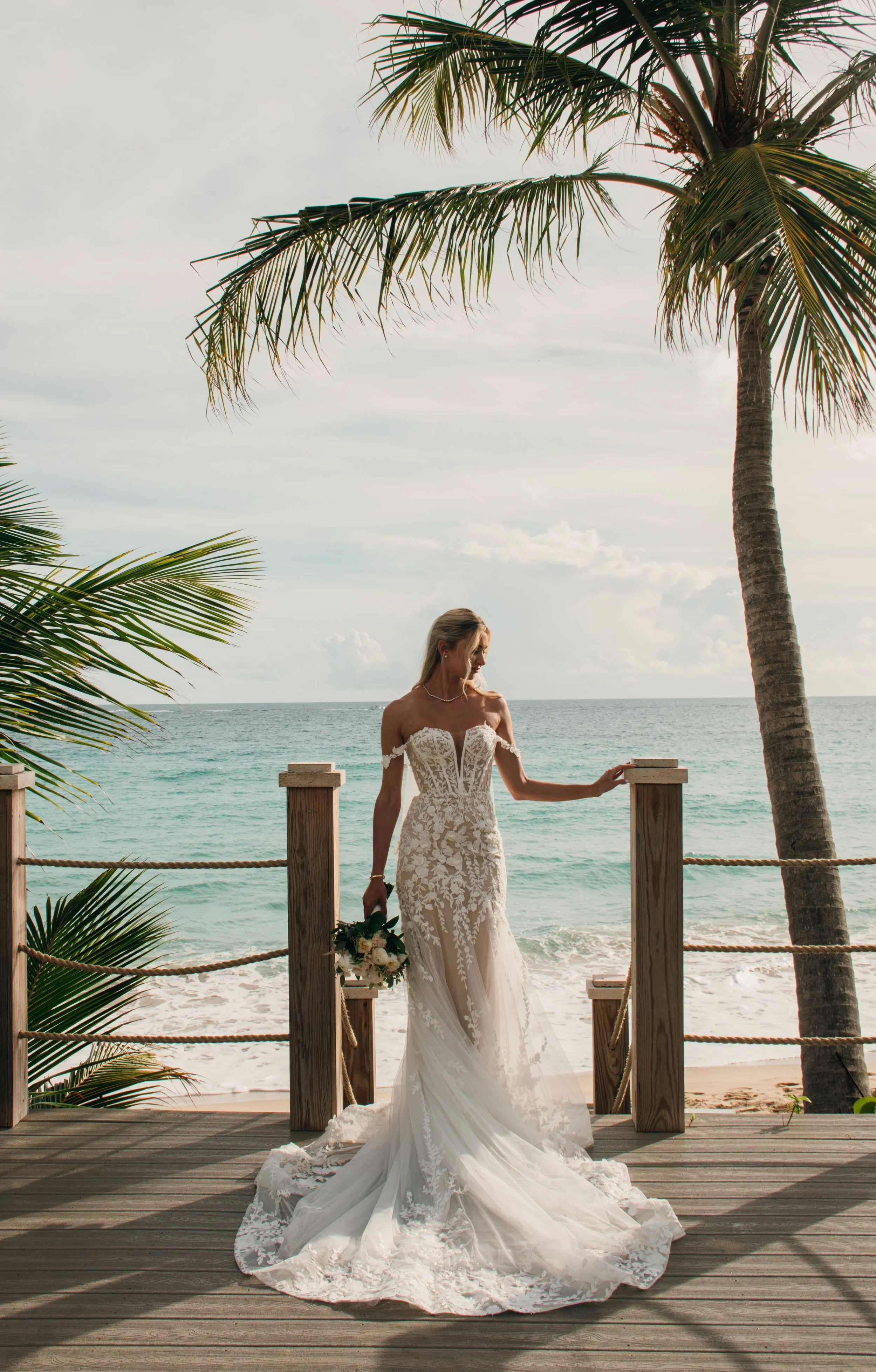 Bride in a white lace wedding dress on a wooden platform overlooking the ocean, surrounded by palm trees.