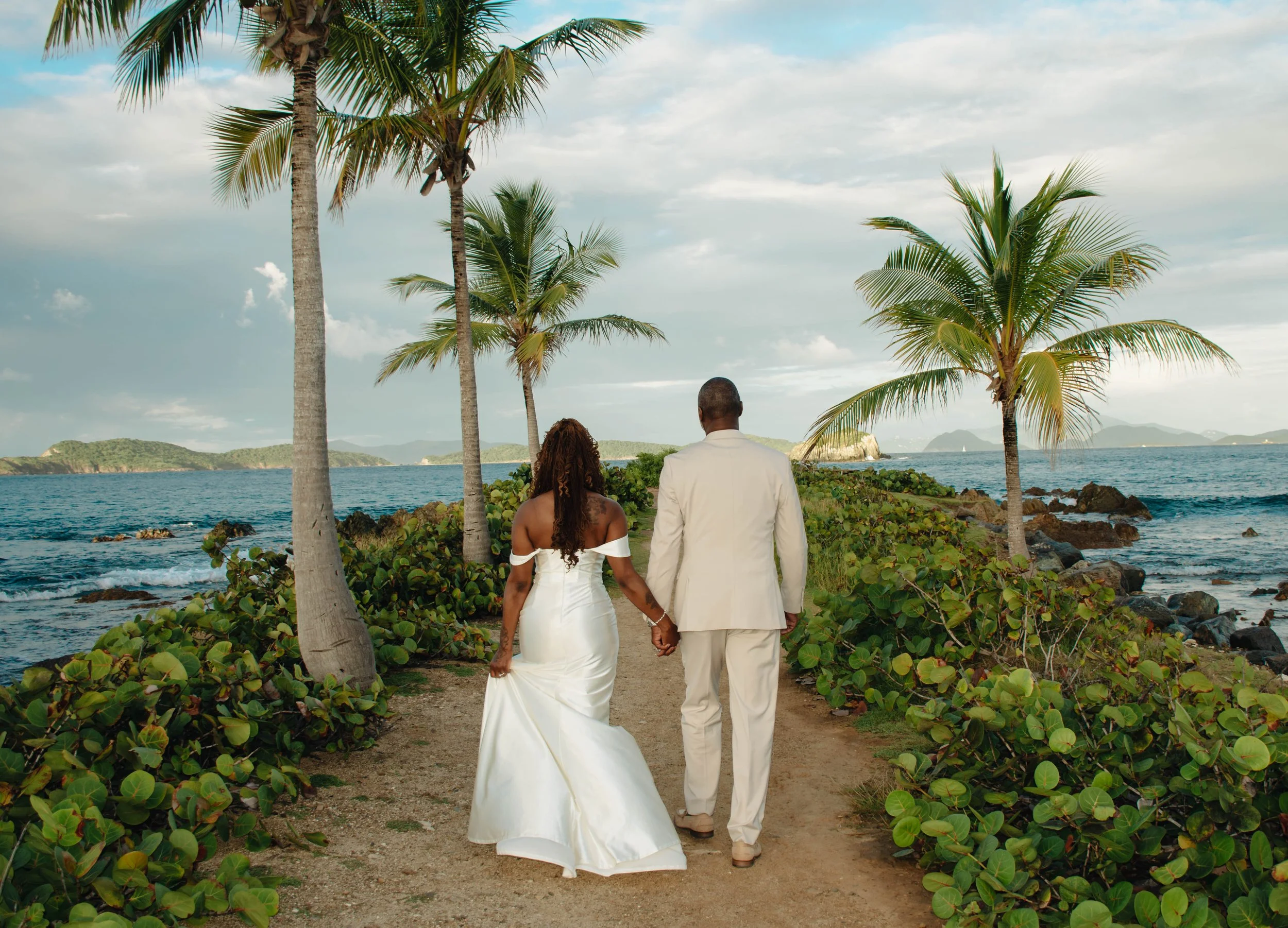 A couple dressed in wedding attire walking hand in hand along a beach path with palm trees, ocean, and islands in the background.