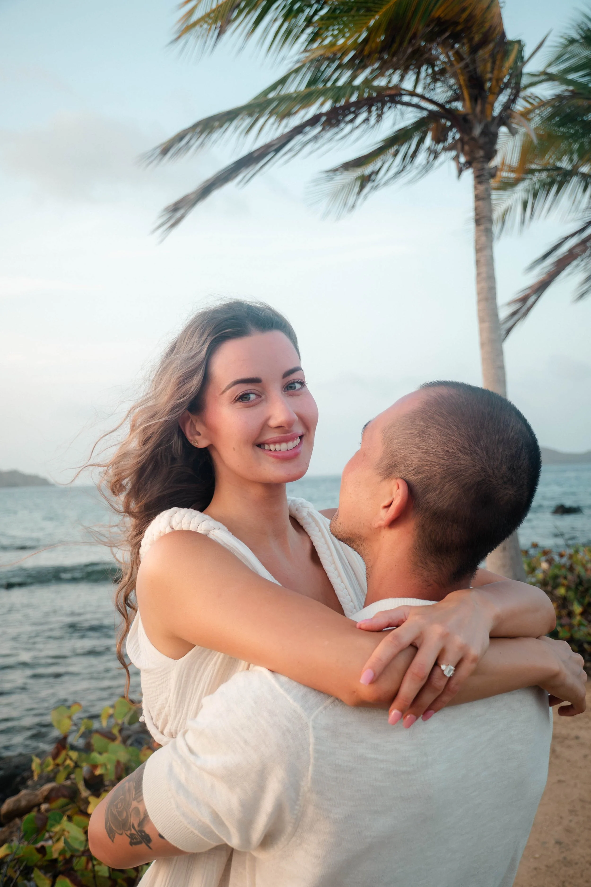 A smiling woman with long, wavy hair embracing a man near a beach, with palm trees and water in the background.
