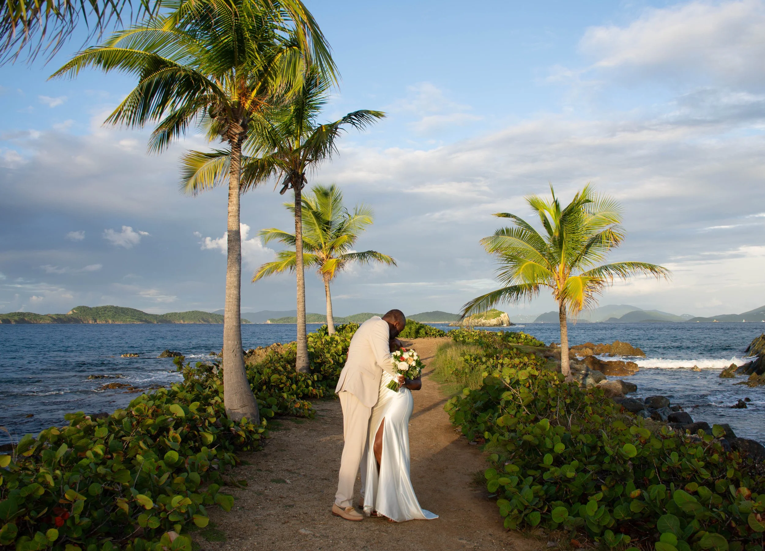 A couple dressed in wedding attire, holding a bouquet, standing on a tropical beach with palm trees, water, and distant islands under a partly cloudy sky.