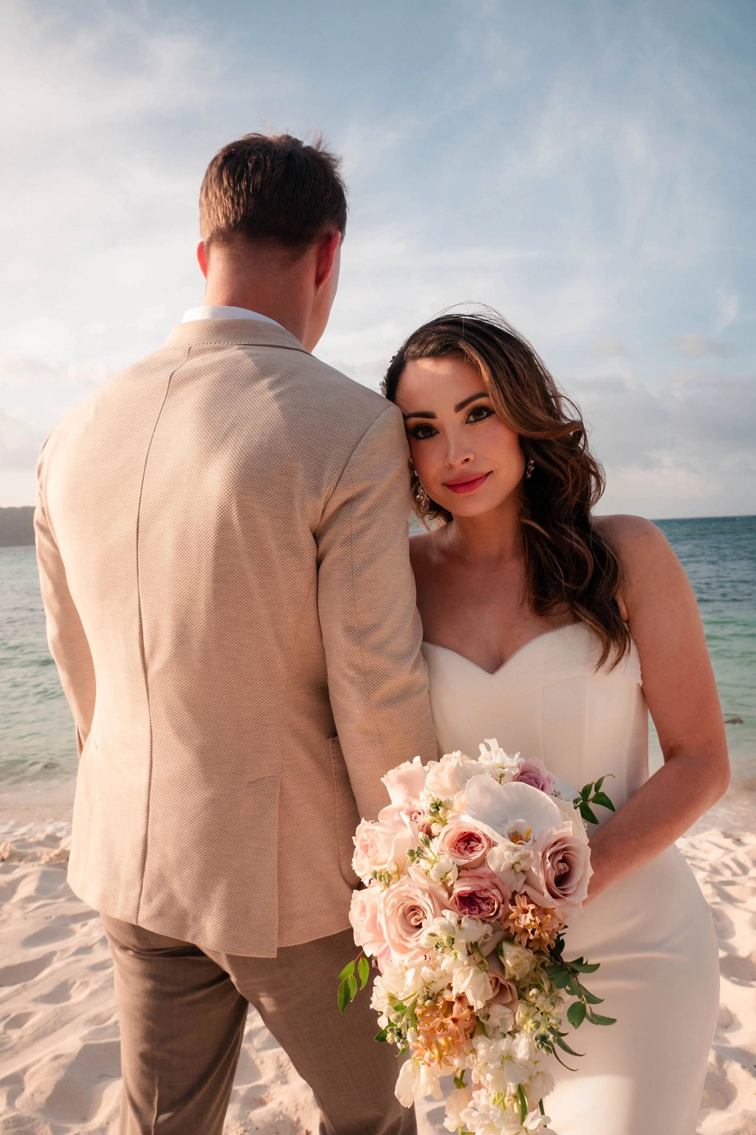 A bride and groom stand close together on a beach, with the groom facing away and the bride holding a large cascading bouquet of pink and white flowers. The bride looks at the camera with a gentle smile, and the sky is partly cloudy in the background