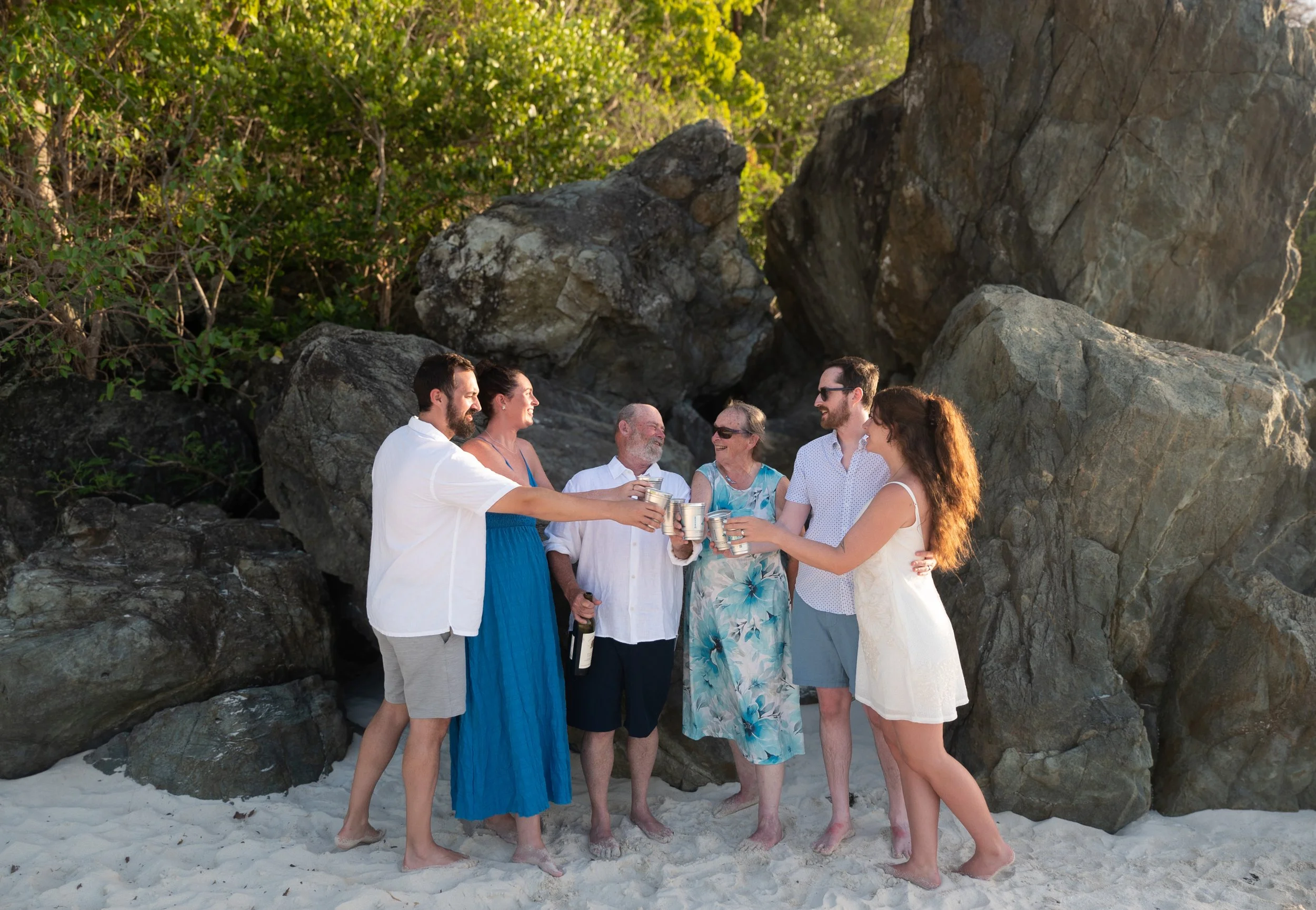 A group of six people celebrating with drinks on a beach in front of large rocks and green foliage.