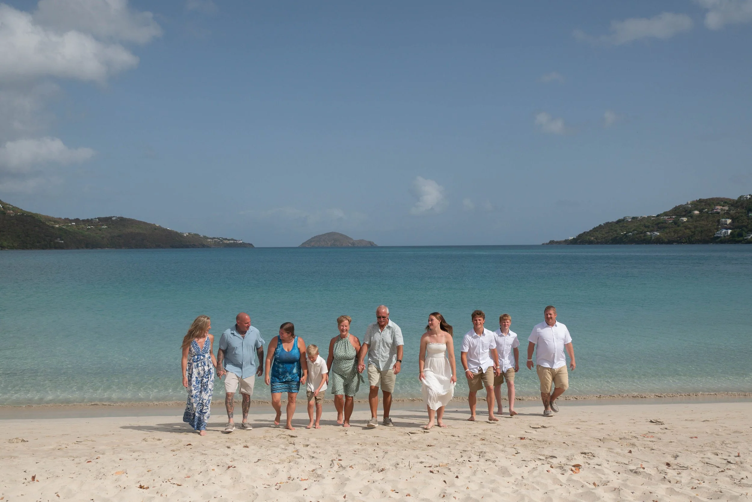 A group of people walking along a beach shoreline with the ocean, hills, and a small island in the background on a sunny day.