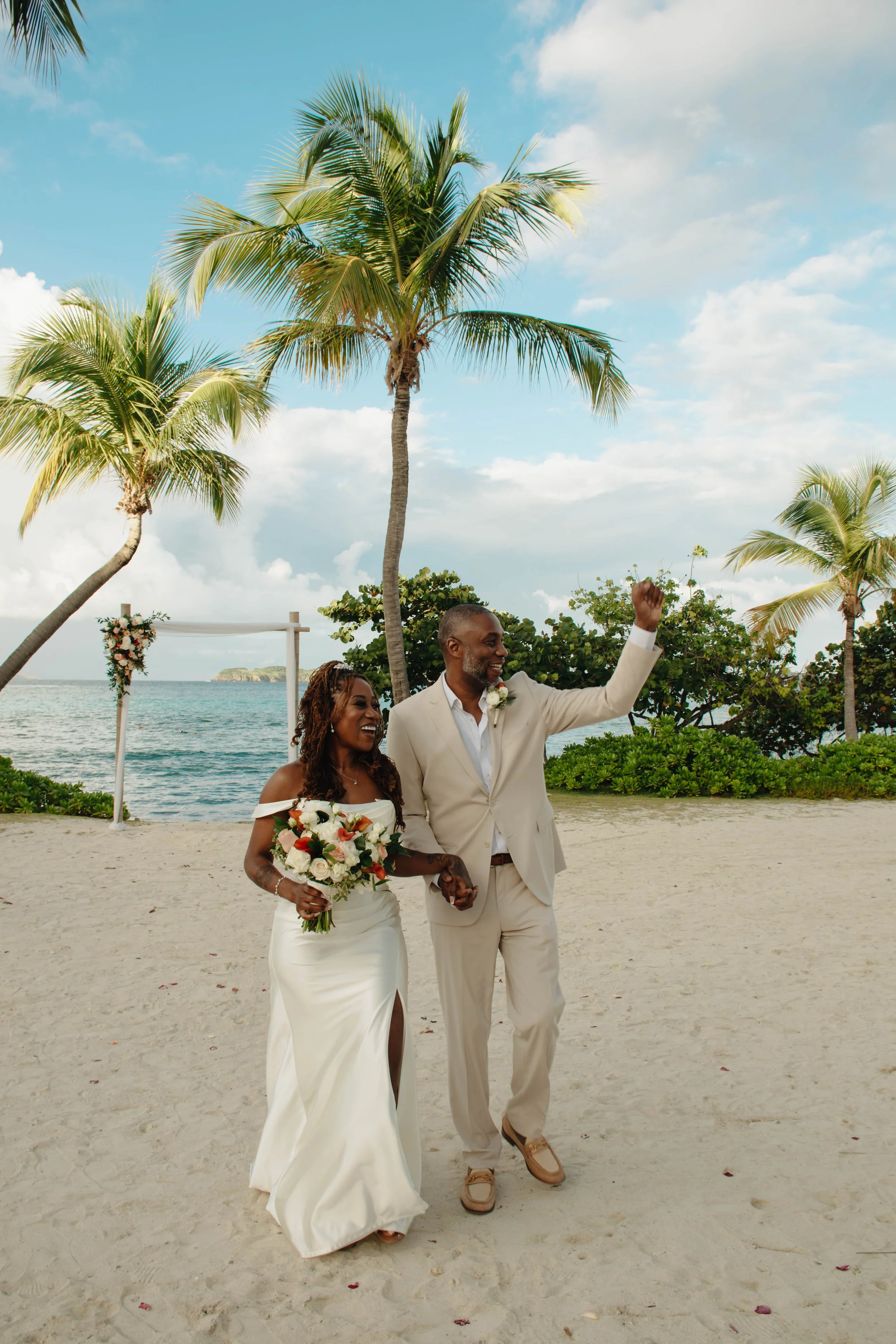 A newly married couple walking on a beach, holding hands, with palm trees and an ocean in the background. The bride is wearing a white gown and holding a bouquet of flowers. The groom is dressed in a beige suit and raising his fist in celebration.