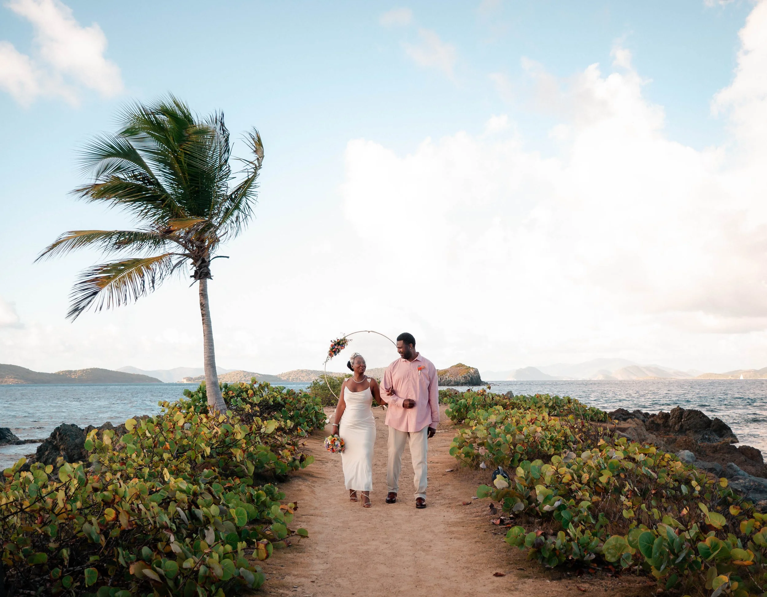 A couple is walking hand in hand along a sandy beach with greenery and rocks. The woman is wearing a white dress and holding a bouquet, while the man is dressed in a pink shirt and light-colored pants. There is a tall palm tree, ocean, and mountains 